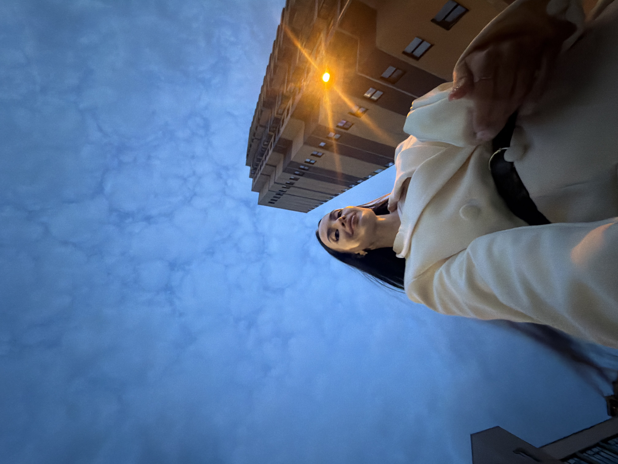 A low-angle, wide-shot photograph captures a woman in a cream-colored blazer and pants standing outside at dusk. She is looking up and slightly to her left, with a subtle smile. Behind her, a tall, multi-story apartment building with a light orange facade and dark brown accents dominates the background, its windows illuminated by a warm streetlamp. The sky above is a deep blue, textured with wispy clouds, indicating a twilight setting. The foreground shows the woman's shoulder and chest, with her right hand reaching up towards her chest, her fingers lightly touching the fabric of her blazer. The overall impression is a candid, urban moment captured as the day transitions into night.