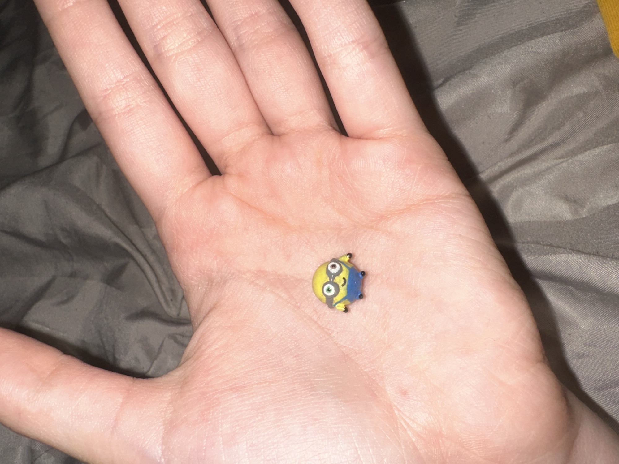 A close-up, overhead shot shows a person's open palm holding a tiny yellow toy minion. The minion is round with black goggles, blue overalls, and black shoes. The palm is light-skinned with visible lines and texture. The background is a dark gray, wrinkled fabric, possibly a blanket or sheet. There is no identifiable context for the location.