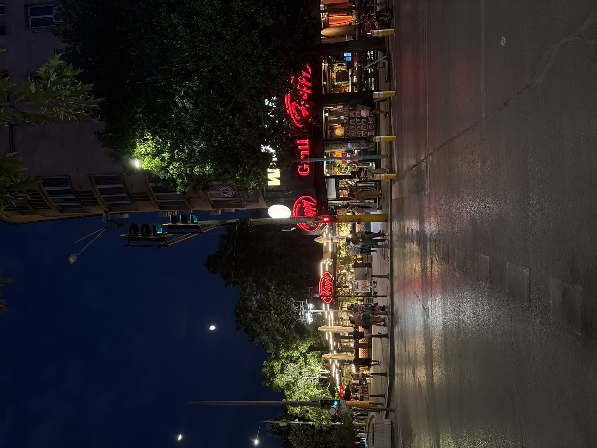 The image captures a street scene at night in Sofia, Bulgaria, likely in an area with commercial establishments and pedestrian activity. A prominent neon sign reading "Happy Grill" in red letters is visible on the facade of a building. The street is wet, reflecting the lights from the surrounding businesses and street lamps. Several people are walking on the sidewalk, some in groups. Traffic lights are illuminated, indicating the presence of an intersection. Trees line the street, and the dark night sky features a crescent moon. The architecture suggests an urban environment with multi-story buildings. The overall atmosphere is that of a lively evening in the city.
