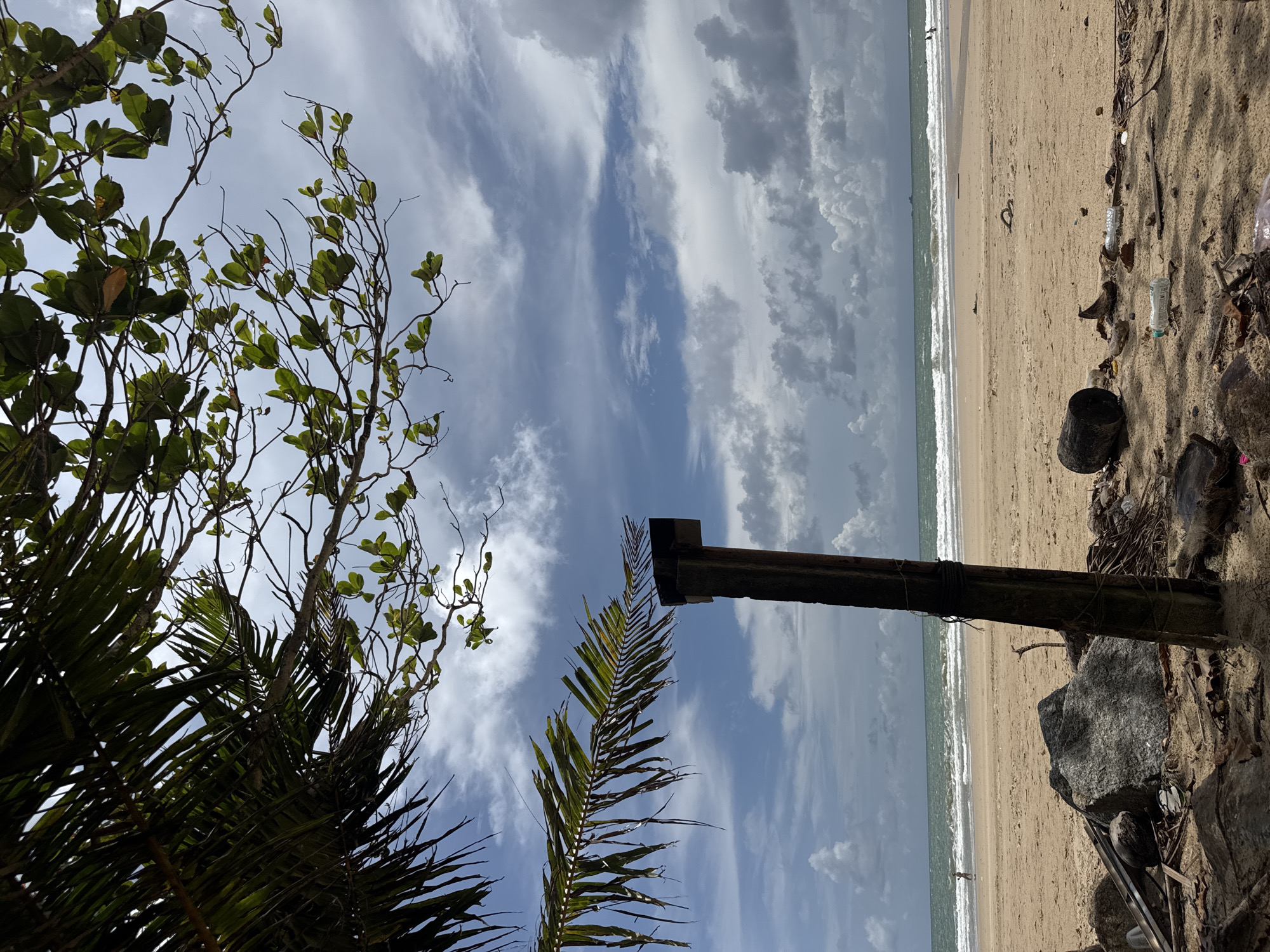 This is a low-angle, full outdoor shot of a beach in Choeng Thale, Thailand, under a partly cloudy sky. In the foreground, palm fronds and tree branches are visible, obscuring the upper portion of the sky. A weathered wooden post, secured with rope, stands prominently in the center of the frame. The beach itself is sandy, with scattered rocks and debris, including plastic bottles, coconut husks, and driftwood. The ocean is visible in the background, with moderate waves breaking on the shore. A single person is walking on the beach in the distance. The overall scene suggests a natural environment impacted by human presence.