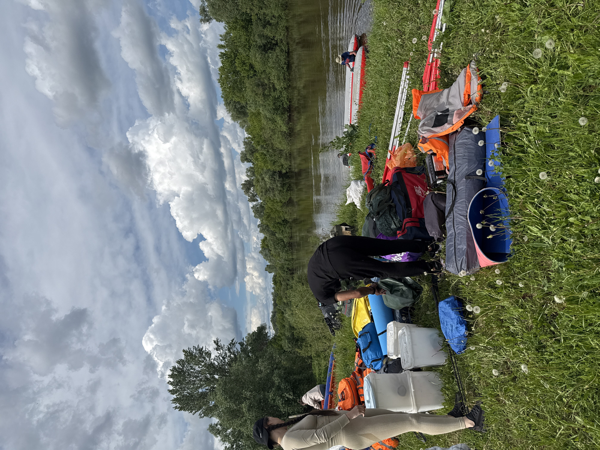 The scene depicts a riverbank in Nazyar, Russia, with people preparing for a river trip. An inflatable kayak is partially in the water, occupied by a person wearing a hat and dark clothing. On the grassy bank, two other individuals are present. One is wearing a beige outfit and a black cap, standing near a pile of camping equipment, including backpacks, blue sleeping pads, white containers, and orange dry bags. Another person is bent over the gear, appearing to be sorting or packing it. The background features a river surrounded by lush green trees under a sky filled with dramatic cumulus clouds. Dandelions are visible in the foreground grass.
