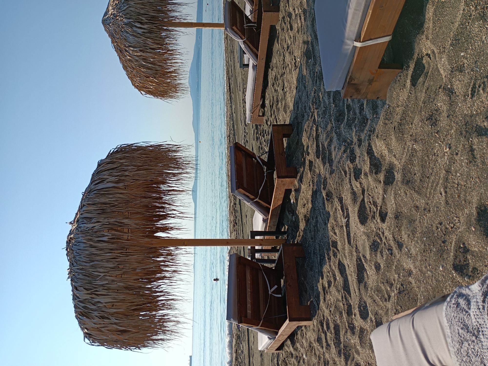 The image depicts a serene beach scene on a sunny day. The foreground is dominated by dark, coarse sand, with several wooden lounge chairs with grey cushions arranged facing the sea. Two large, thatched umbrellas provide shade over some of the chairs. In the middle ground, the calm, blue sea gently laps at the shore, and a person is visible swimming. The background features a hazy coastline with mountains in the distance, and a clear blue sky overhead. The overall impression is one of relaxation and tranquility. This scene is located on the beach in Burgas, Bulgaria.