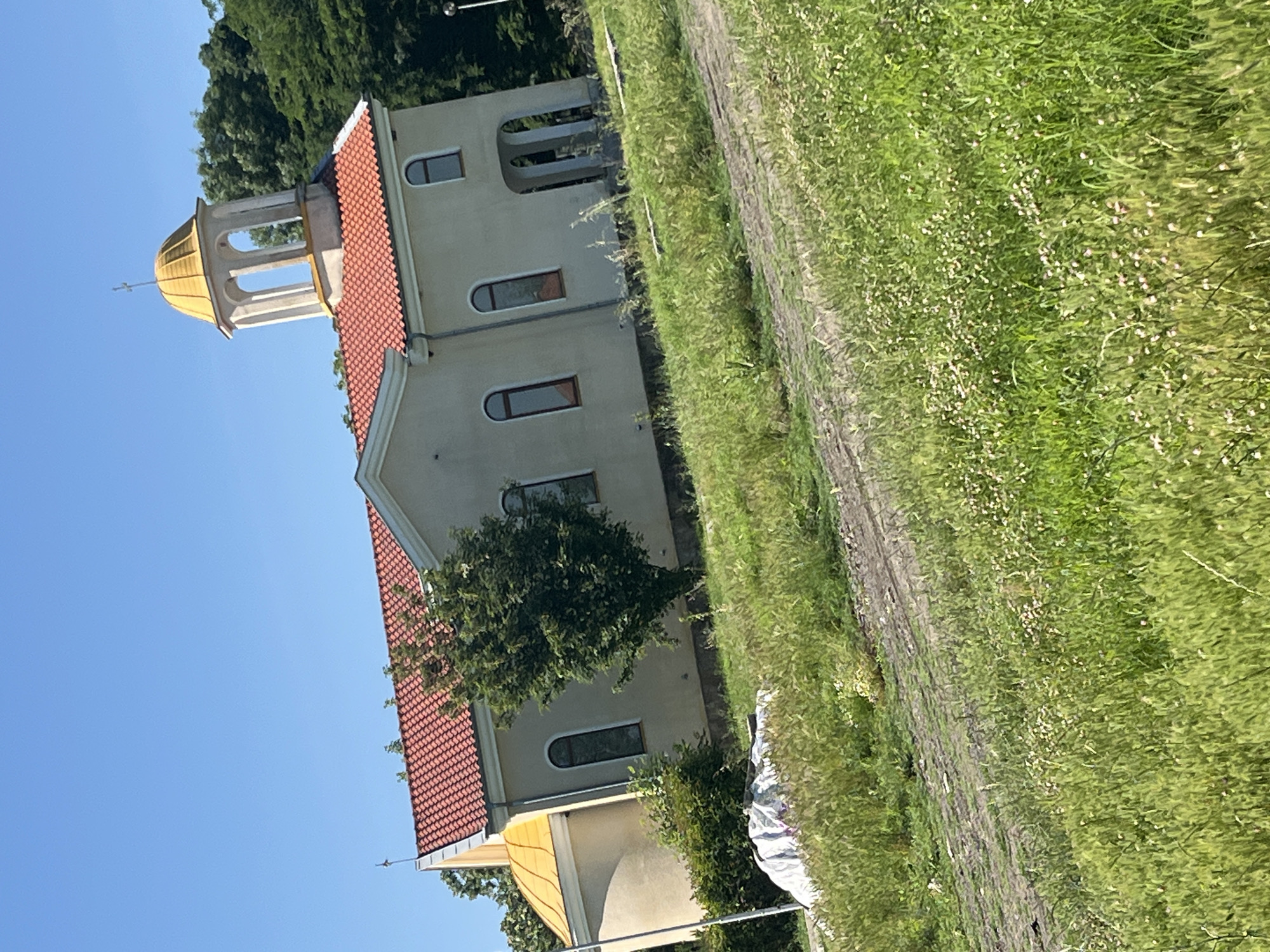 The image depicts a church located in the village of Ravnets, Bulgaria. The church has a light-colored facade, a red tiled roof, and a yellow dome. A bell tower with a golden cross is visible on top of the bell tower. The church is surrounded by a grassy field with some trees in the background. A dirt path runs through the field in front of the church. There are no people visible in the image. The scene is illuminated by bright sunlight, indicating it is daytime.