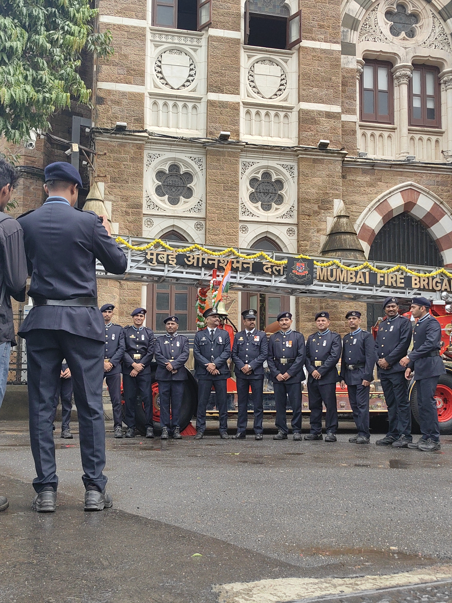 The image captures a group of eight men in dark blue uniforms, likely firefighters, standing in formation in front of a red fire truck. They are positioned on a wet, paved street with puddles, suggesting recent rain. Behind them is a large, ornate brick building with arched windows and decorative white stonework, indicative of colonial-era architecture. A banner above the fire truck reads "MUMBAI FIRE BRIGADE" in English, with accompanying text in Marathi script, confirming the location as Mumbai, India. The men are formally dressed, with some wearing berets and others peaked caps. The overall scene appears to be a ceremony or official gathering.