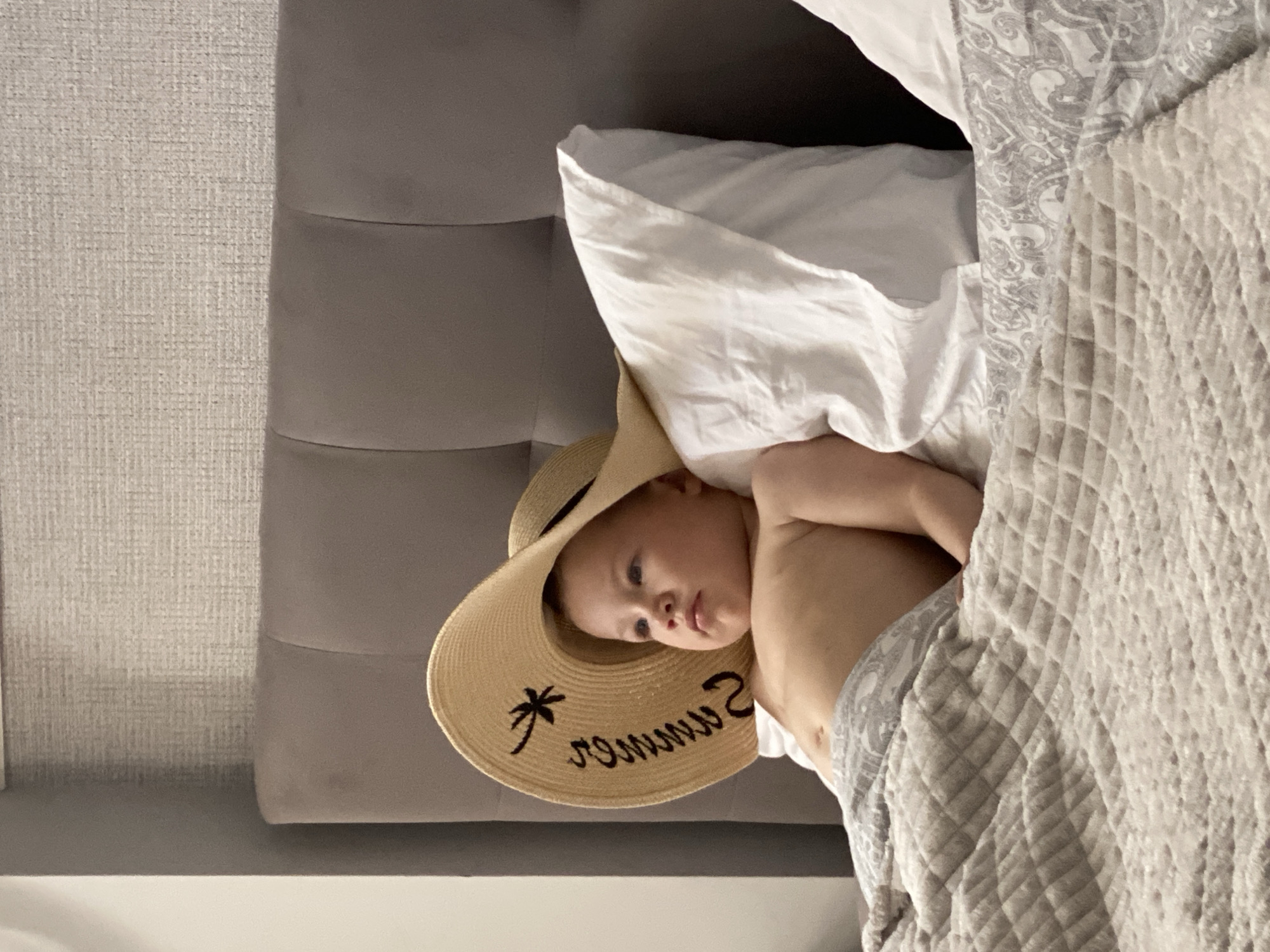 An infant wearing a large straw hat is lying in a bed. The hat has "SUMMER" written on it in black cursive lettering, along with a palm tree graphic. The infant is lying on their back, propped up by a white pillow, and is partially covered by a quilted grey blanket with a paisley pattern. The bed is in a room with a grey headboard and textured wallpaper. The background is blurred, but appears to be an indoor setting.