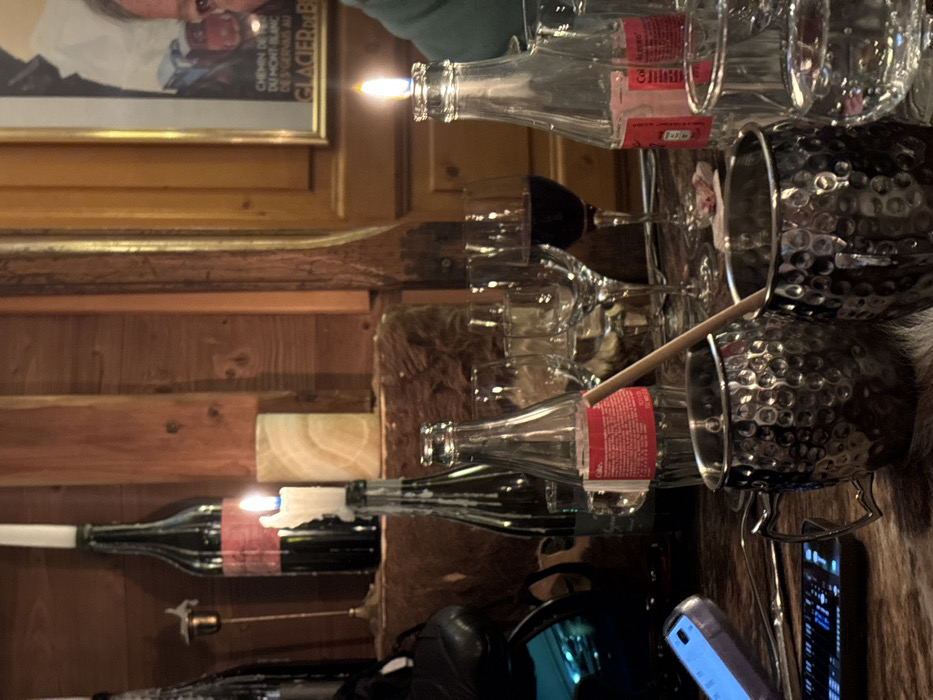 This image captures a cozy, rustic indoor scene, likely in a restaurant or bar in Val-d'Isère, France, during the evening. The main subjects are an assortment of glassware and bottles arranged on a surface, possibly a wooden bar or table, partially covered by a furry hide.
In the foreground and midground, several empty clear glass bottles are visible, one with a red label. Two bottles serve as makeshift candle holders, with lit candles emanating a warm glow, and melted wax dripping down a dark green wine bottle. A collection of empty wine glasses and tumblers are stacked and placed nearby. To the right, two metallic, hammered-texture mugs are seen within a metal caddy, one containing a wooden stirring stick. A smartphone displaying information lies on the right side of the frame, alongside a dark, reflective object.
The background features warm, rustic wooden paneling, typical of an alpine chalet. A framed picture hangs on the wall, showing a close-up of a man's smiling face. Visible text on the picture includes "CHEMIN DE FER DU MONT BLANC DES SERVOZ AU" and "GLACIER DE BLANCS", clearly indicating a connection to the Mont Blanc region in the French Alps, reinforcing the location context.
The scene suggests a relaxed gathering or dining experience, highlighted by the use of candlelight for ambiance. The lighting indicates it is nighttime or a dimly lit interior. No people are actively present in the immediate scene, only their traces in the form of used glassware. The overall atmosphere is intimate and comfortable, characteristic of a mountain resort establishment.