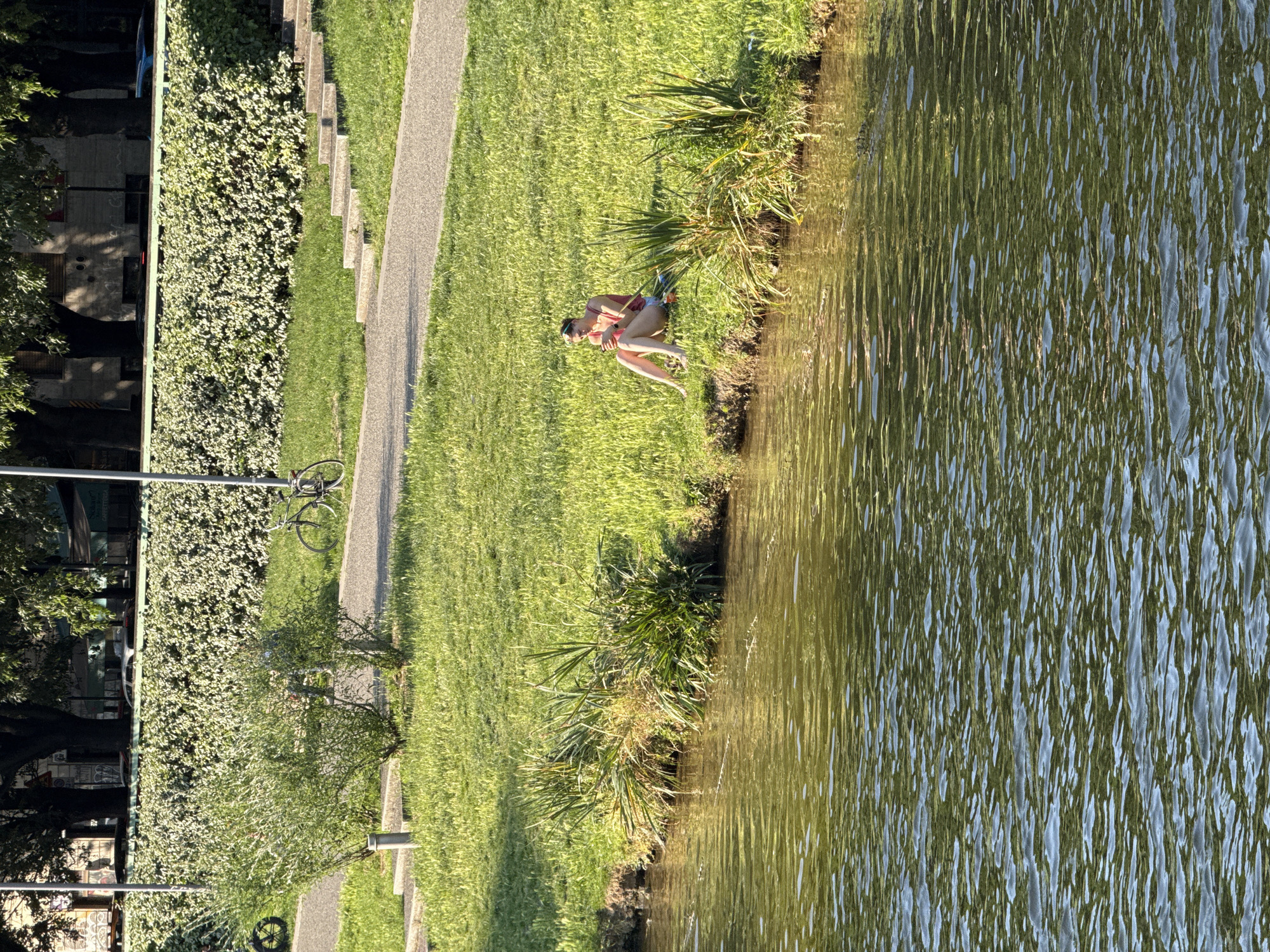 A landscape image shows a woman sitting on a grassy riverbank, likely in Milan, Italy. The woman is wearing a swimsuit. The river's water surface is textured with ripples. A paved pathway runs parallel to the riverbank, lined with vegetation and trees. A bicycle stands next to a street light pole. In the background, buildings and more trees line the landscape.