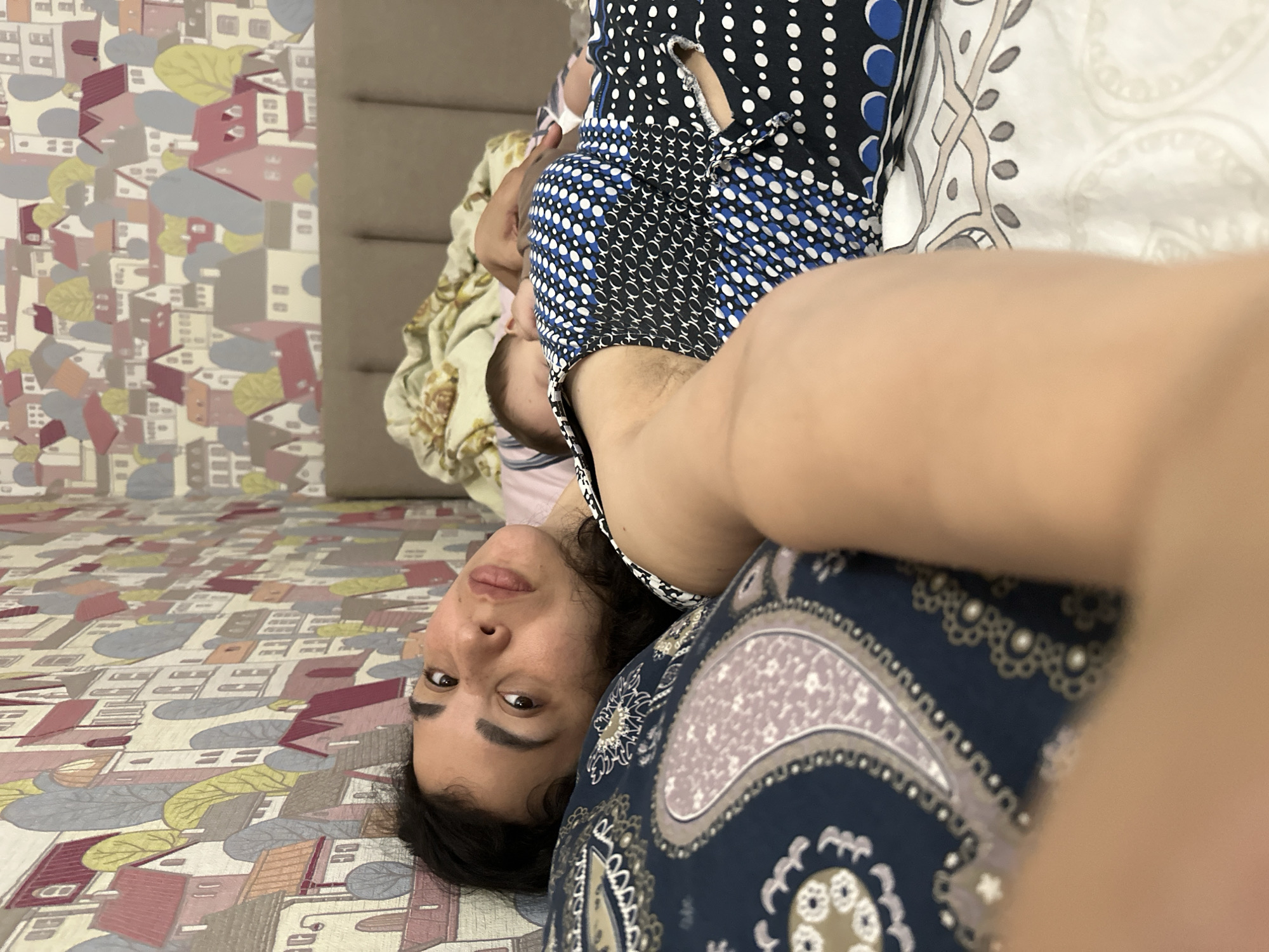 An indoor scene captured from a close perspective shows an adult and a partially visible child, oriented sideways due to the image being rotated 90 degrees clockwise.
The main subject is an adult, likely female, lying down on her back with her head towards the bottom left of the rotated frame, looking directly into the camera. She has dark hair, dark eyes, and a neutral to slightly content expression. She is wearing a patterned garment that appears to be a dress or top, featuring sections of black with white polka dots, blue with white polka dots, and blue with a white circular/chain-link pattern. A smaller portion of a black and white patterned strap or garment is also visible near her shoulder. Her arm is extended towards the camera, suggesting a selfie. She is also partly covered by or wearing a bottom garment with a dark blue background and a complex paisley-like pattern in lighter shades of blue, grey, and cream/tan.
Behind the adult's head, a baby or small child is partially visible, appearing to be lying next to her or in her arms. The child's head and a small portion of a light-colored, possibly floral-patterned blanket or clothing are visible.
The surface beneath the subjects, which would be the bed or floor, is covered with a distinctive patterned textile. This pattern depicts an aerial or stylized view of a cityscape, featuring numerous houses, buildings, and trees in muted colors such as pale red, grey, cream, yellow, and blue.
In the upper right background of the rotated image, a plain, light brown padded headboard or piece of furniture is visible. To the right of the adult subject, a section of a white or cream-colored sheet or pillow with a faint, intricate pattern can be seen. No visible text is present in the image.