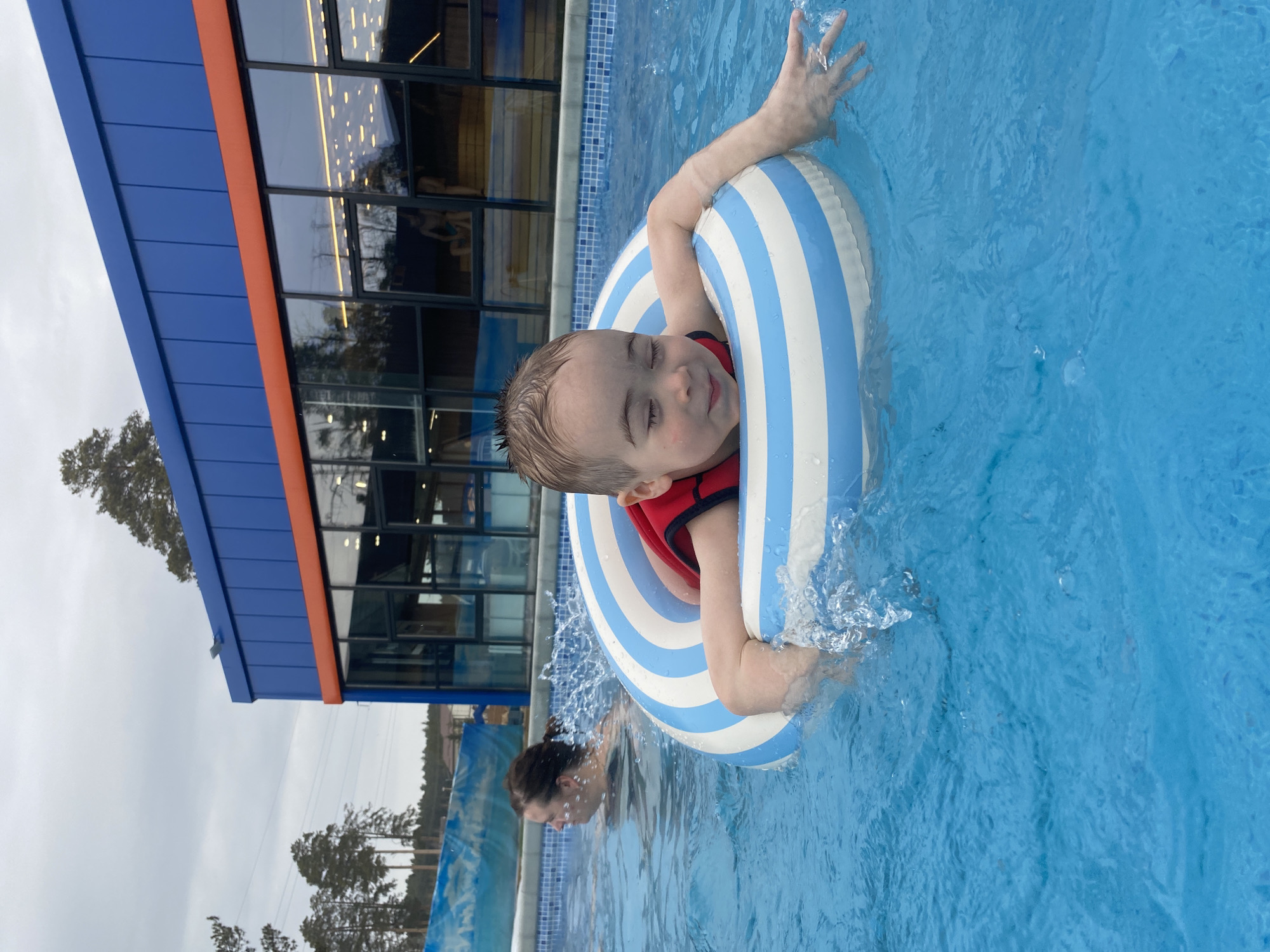 An outdoor swimming pool scene in the city of Votkinsk, Russia, is depicted on an overcast day. In the foreground, a young male child, with wet light brown hair, floats in a white and light blue horizontally striped inflatable ring. He wears a red life vest, his eyes are closed, and he displays a slight, relaxed smile. His arms are draped over the ring, with his right hand extended, creating small splashes in the water. To the left and slightly behind, an adult female with dark hair tied back in a ponytail is partially submerged, facing left, with water splashing around her shoulders.
The pool water is bright blue, and a tiled edge with a small blue and white mosaic pattern is visible. In the background, a modern two-story building features a dark blue panelled upper facade and a lower section comprising large, dark-framed rectangular windows, segmented by a prominent orange horizontal band. The windows reflect interior lighting and a single individual, possibly male, seated within. Beyond the building and to the left, several tall pine trees are visible against the grey, cloudy sky. Faint power lines traverse the distant horizon amidst the trees. A blue structure with a wave-like pattern is partially visible to the far left of the adult swimmer.
