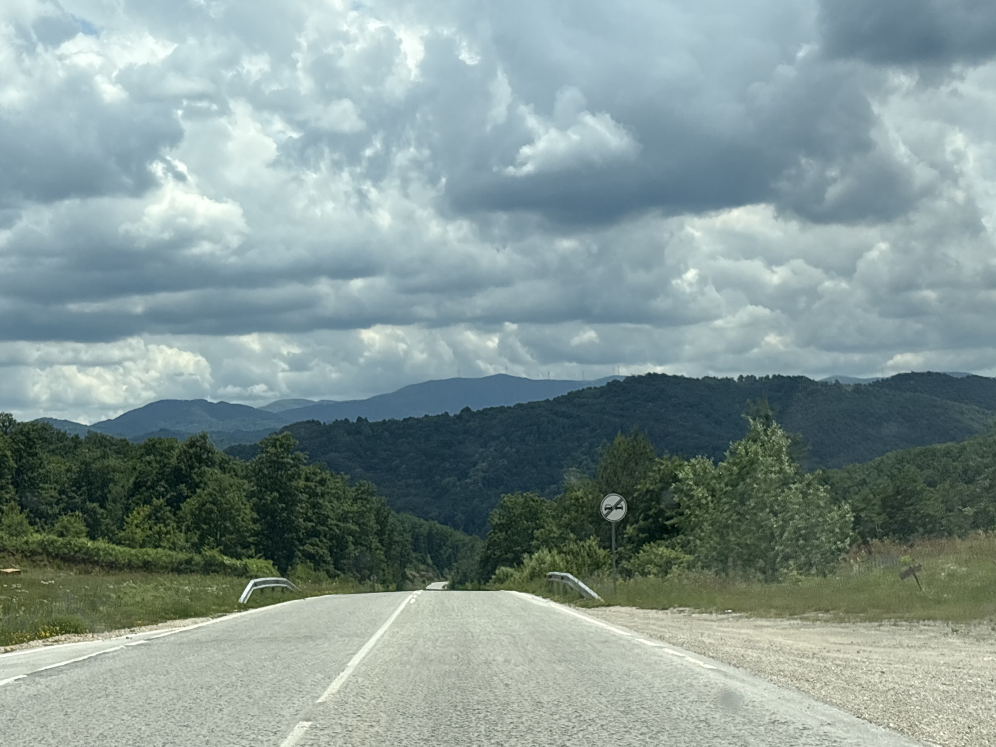 The image depicts a two-lane asphalt road in a rural, mountainous region under a cloudy sky. The road has a white dashed line separating the lanes and a solid white line on the right edge. Guardrails are visible on both sides of the road. To the right, there is a road sign with a circular white background and a red border, featuring a black symbol of a horn with a diagonal line through it, indicating no honking. The landscape features lush green trees and hills, with more distant, darker-hued mountains in the background. The overall impression is of a scenic drive on a potentially overcast day. This view is consistent with a road leading into or out of the city of Gabrovo, Bulgaria, which is known for its surrounding mountainous terrain.
