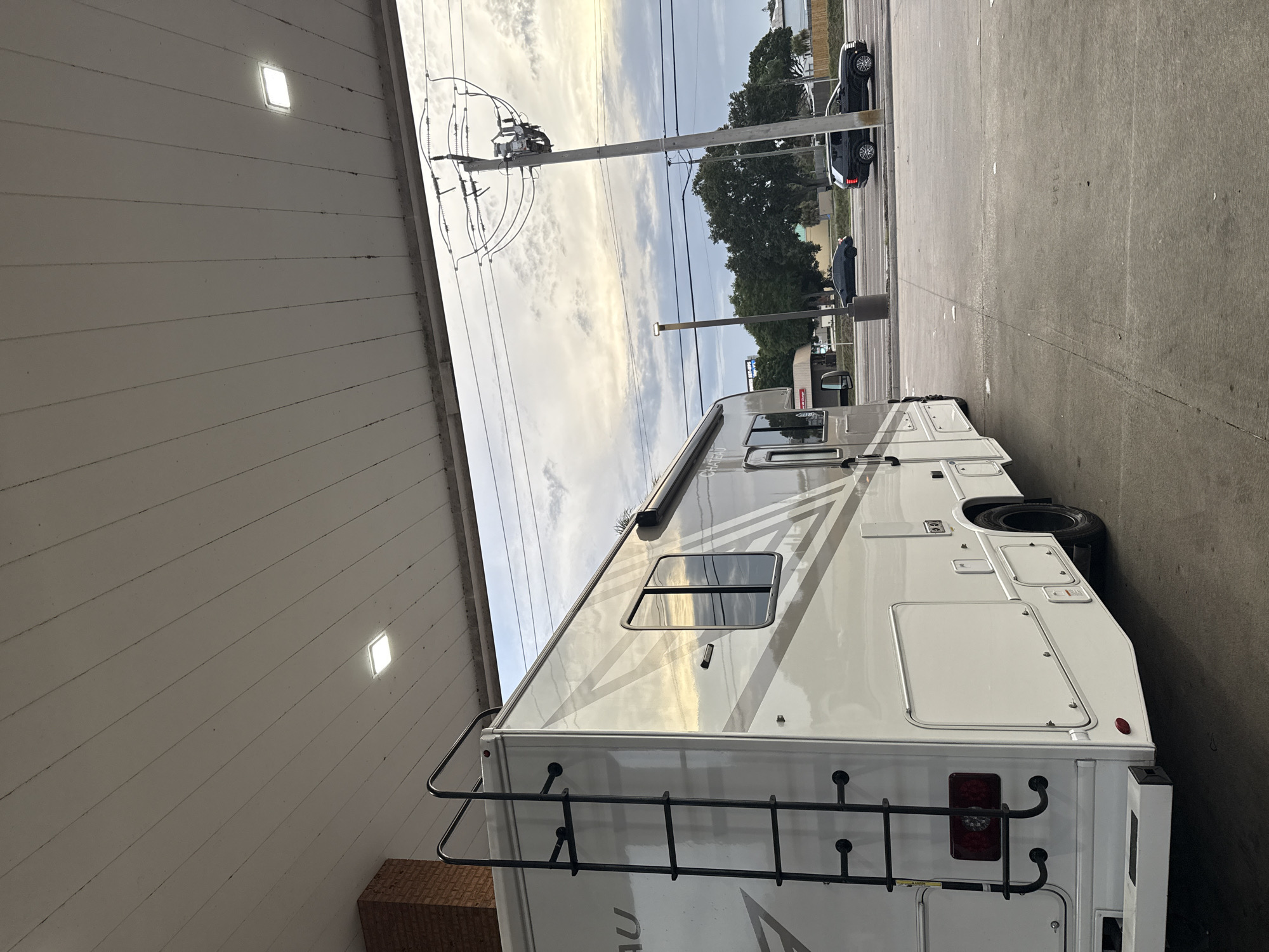 The image depicts a white motorhome parked at a gas station. The scene is captured during daylight hours under a cloudy sky. The motorhome, featuring a ladder at the rear, is positioned near a gas pump under a gas station canopy. Power lines and poles are visible in the background. A dark-colored vehicle is parked beyond the motorhome, along with trees and street lamps. Due to the lack of specific visual identifiers like street signs, or landmarks, pinpointing the exact location within Palm Bay, Florida, is not possible.