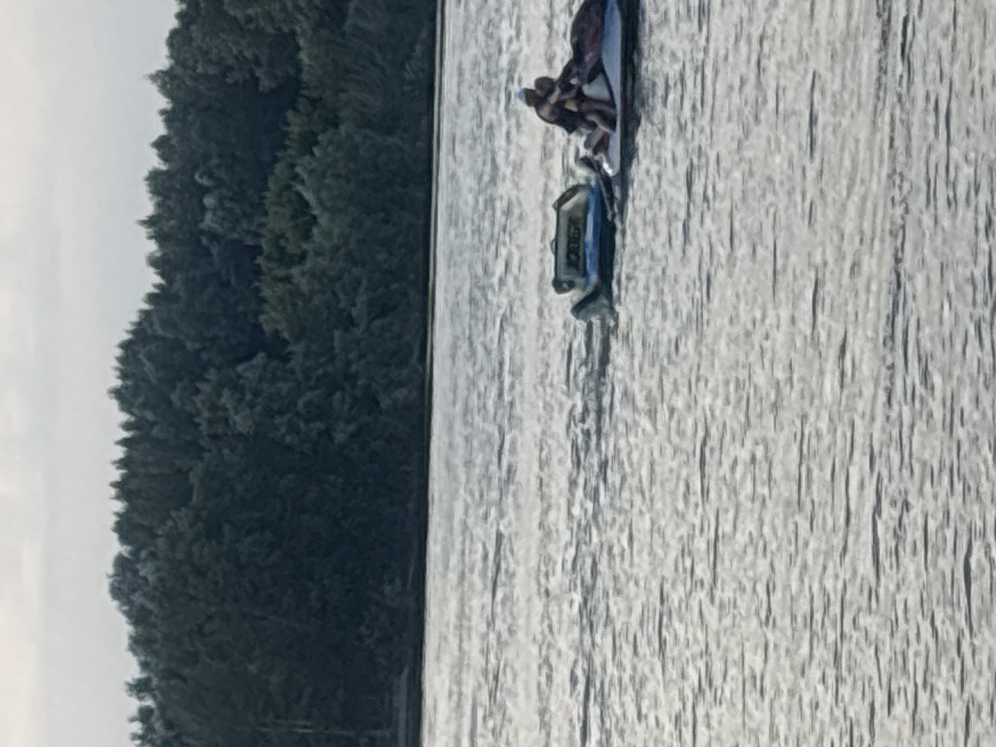 Two individuals are on a jet ski in the Irtysh River, near the shore of Pavlodar, Kazakhstan. The jet ski is white and red, and the two people are wearing dark clothing. Behind them, a blue and yellow inflatable towable tube is drifting in the water. The river is calm, with ripples reflecting the overcast sky. In the background, a dense line of green trees lines the riverbank. The overall scene depicts a recreational activity on a body of water.