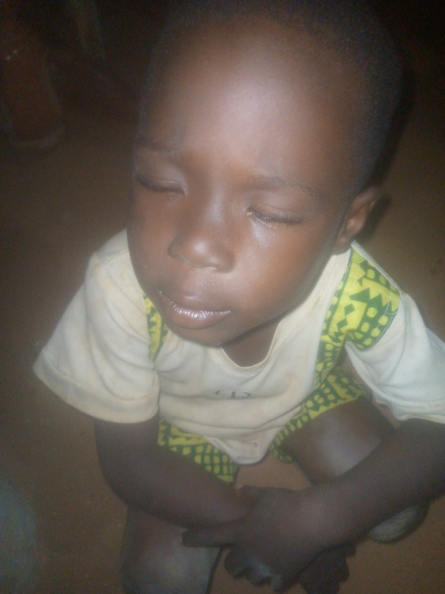 A young, dark-skinned child is seated indoors. The child has closed eyes with tears visible, suggesting distress. The child is wearing a light-colored shirt with a yellow and green patterned design on the straps or yoke. The child's arms are crossed and close to the body. The background is dark, suggesting a dimly lit room. The location is Unknown, Unknown.