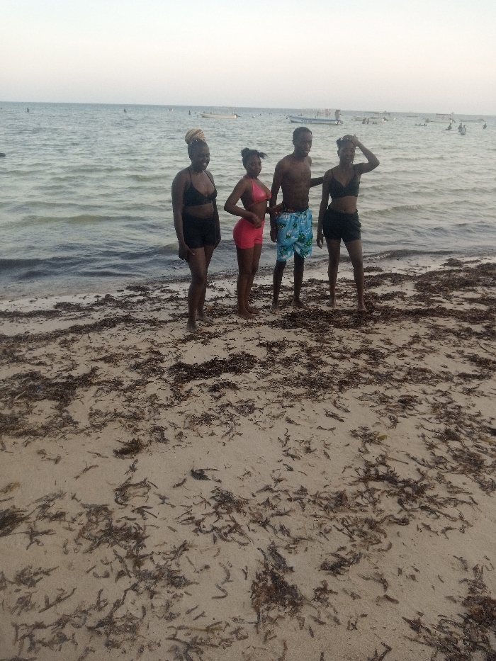 The image depicts a daytime scene on a sandy beach in Shanzu ward, Kenya. Four individuals are positioned in the midground, standing barefoot on the sand. From left to right, the first individual is a female wearing a black bikini top and black shorts, with a light-colored head covering. The second individual is a female in a pink two-piece swimsuit, consisting of a bikini top and shorts, with her left hand resting on her hip. The third individual is a male, shirtless, wearing light blue patterned swim shorts. The fourth individual is a female wearing a black bikini top and black shorts, with her right hand raised to her forehead. All four individuals are facing the camera. The foreground consists of light-colored sand heavily interspersed with dark brown organic debris, consistent with dried seaweed or plant matter. In the background, calm ocean water extends to the horizon. Several small boats are visible on the water, along with multiple indistinct figures partially submerged further offshore. The sky above is clear and light blue.