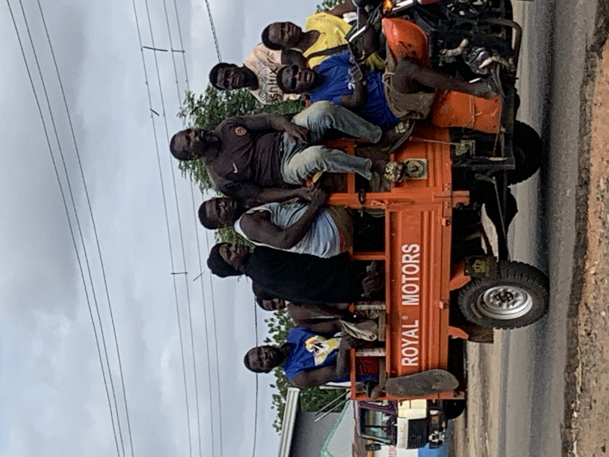 An orange "Royal Motors" branded three-wheeled cargo motorcycle, resembling a motorized tricycle, transports a group of at least nine adult males on a paved road in Shendam, Nigeria. The vehicle is in motion under an overcast sky, with multiple power lines visible horizontally in the upper frame.
Five males are positioned within the open cargo bed. One male on the left wears a blue sleeveless shirt with a white and yellow design. Another male in the center-left wears a black long-sleeved shirt and a black beanie. A third male in the center wears a light blue sleeveless top. A fourth male with a beard, wearing a dark brown short-sleeved shirt and light-colored jeans, sits on the side rail of the cargo bed. A fifth male wears a light-colored "ashlux" t-shirt. Two additional males are partially visible within the cargo bed, behind the first group.
Two males occupy the front motorcycle section. The driver, wearing a blue short-sleeved t-shirt and dark sunglasses, operates the handlebars. A passenger in a yellow t-shirt sits directly in front of the driver, facing forward.
The vehicle displays "ROYAL MOTORS" in white lettering on its orange cargo bed. It features knobby tires and a crowned logo near the rear wheel. A dark-colored mud flap hangs from the left side of the cargo bed. A light-colored container is secured to the frame near the driver's feet.
In the background, a light-colored truck is visible to the left, and a building with a blue-green roof and white wall is further behind it. Green vegetation is present behind the vehicle. The road surface is asphalt, with a visible shoulder or median strip to the right.