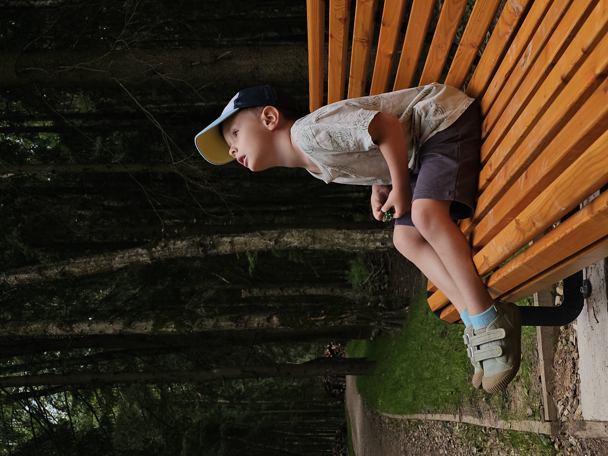 A young boy wearing a baseball cap, t-shirt, and shorts sits on a wooden park bench in a forest setting. He is looking upwards, with a small green object in his right hand. The bench is positioned next to a gravel path, with grass and trees surrounding the area. The trees are tall and dense, suggesting a natural, wooded environment. The lighting indicates it may be daytime, with shadows cast by the trees. The location is identified as Odintsovo, Russia.