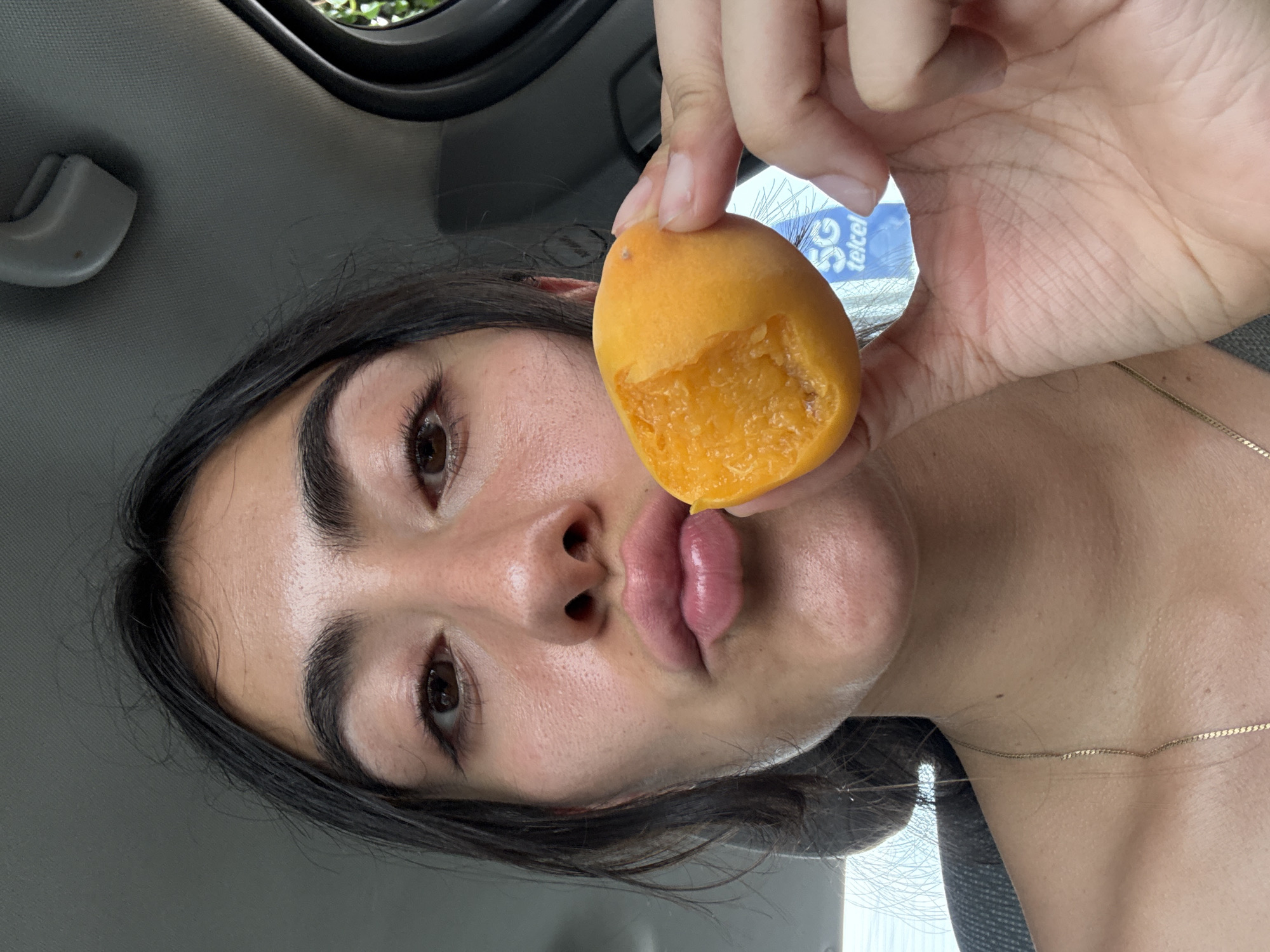 A young woman is holding a partially eaten apricot in front of her face. She is in a car. The background visible through the car window includes foliage and a sign that says “5G telcel.” The location is likely Puebla, Mexico, based on the "telcel" branding which is a Mexican telecommunications company.