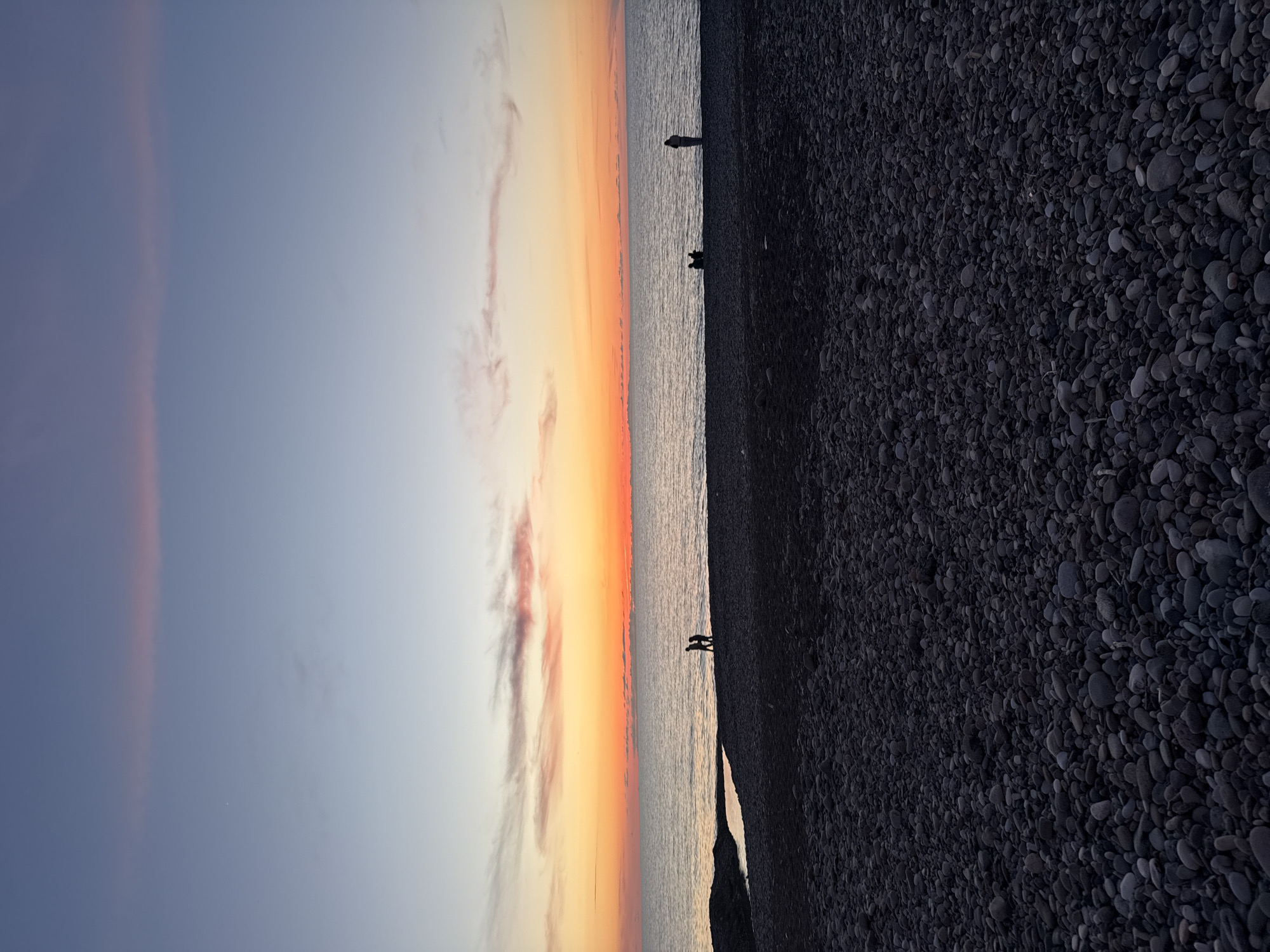 The image depicts a sunset scene at a beach in Batumi, Georgia. The sky exhibits a gradient of colors from light blue and grey to orange and red, indicating the sunset. Three indistinct human silhouettes are visible along the water's edge at varying distances, suggesting people walking or standing on the beach. The beach itself appears to be composed of dark pebbles or stones. The horizon is level with the viewer.