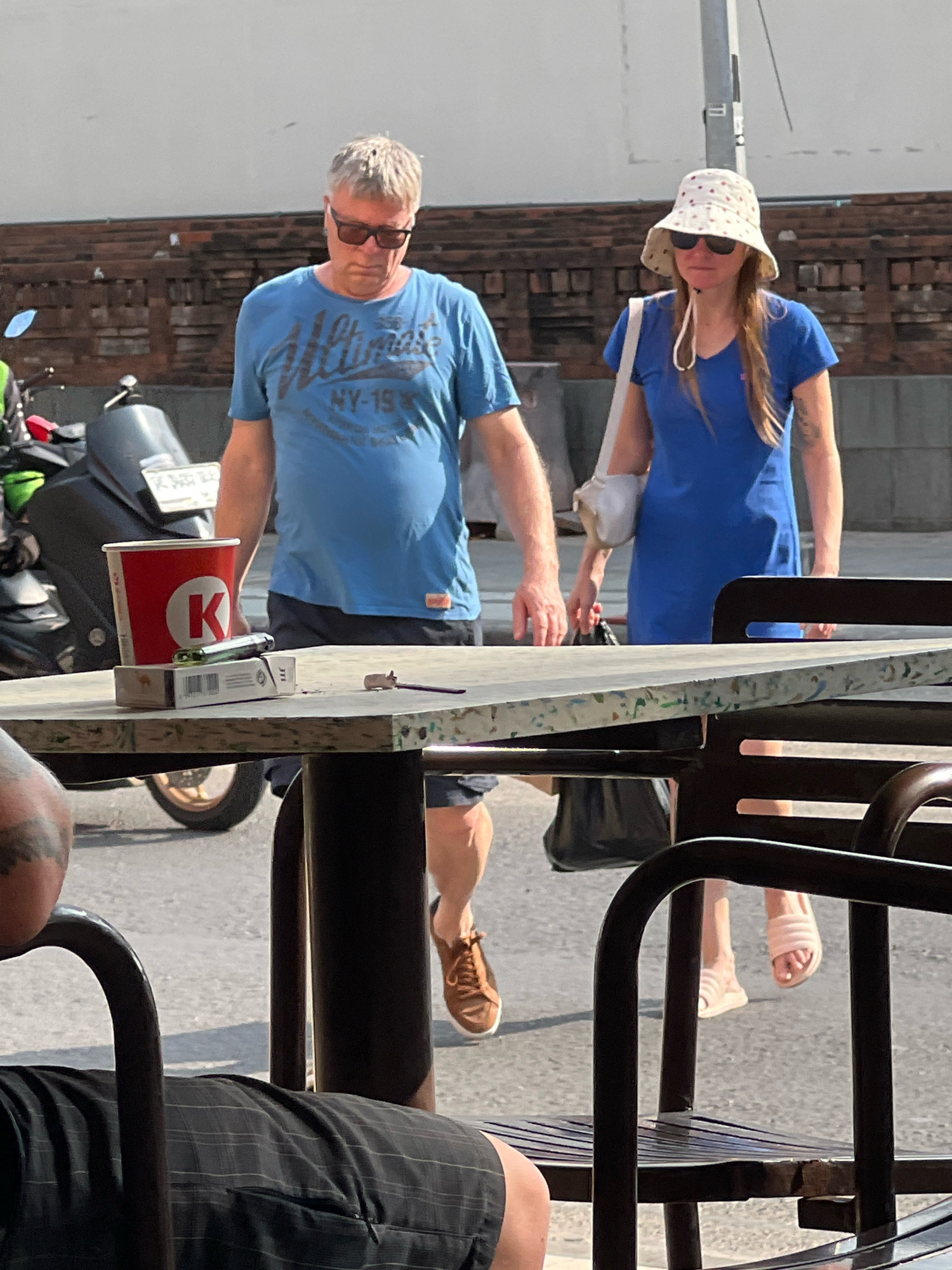 Two individuals, a man and a woman, are walking on a street in Kuta, Indonesia. The man, on the left, wears a blue t-shirt and sunglasses. The woman, on the right, is dressed in a blue dress and a bucket hat, carrying a white bag over her shoulder. They are walking past a table with a red cup, a pack of cigarettes, and a lighter on it. A motorcycle is parked behind the table. The background shows a brick wall and a street. The scene appears to be an outdoor seating area of a cafe or restaurant.