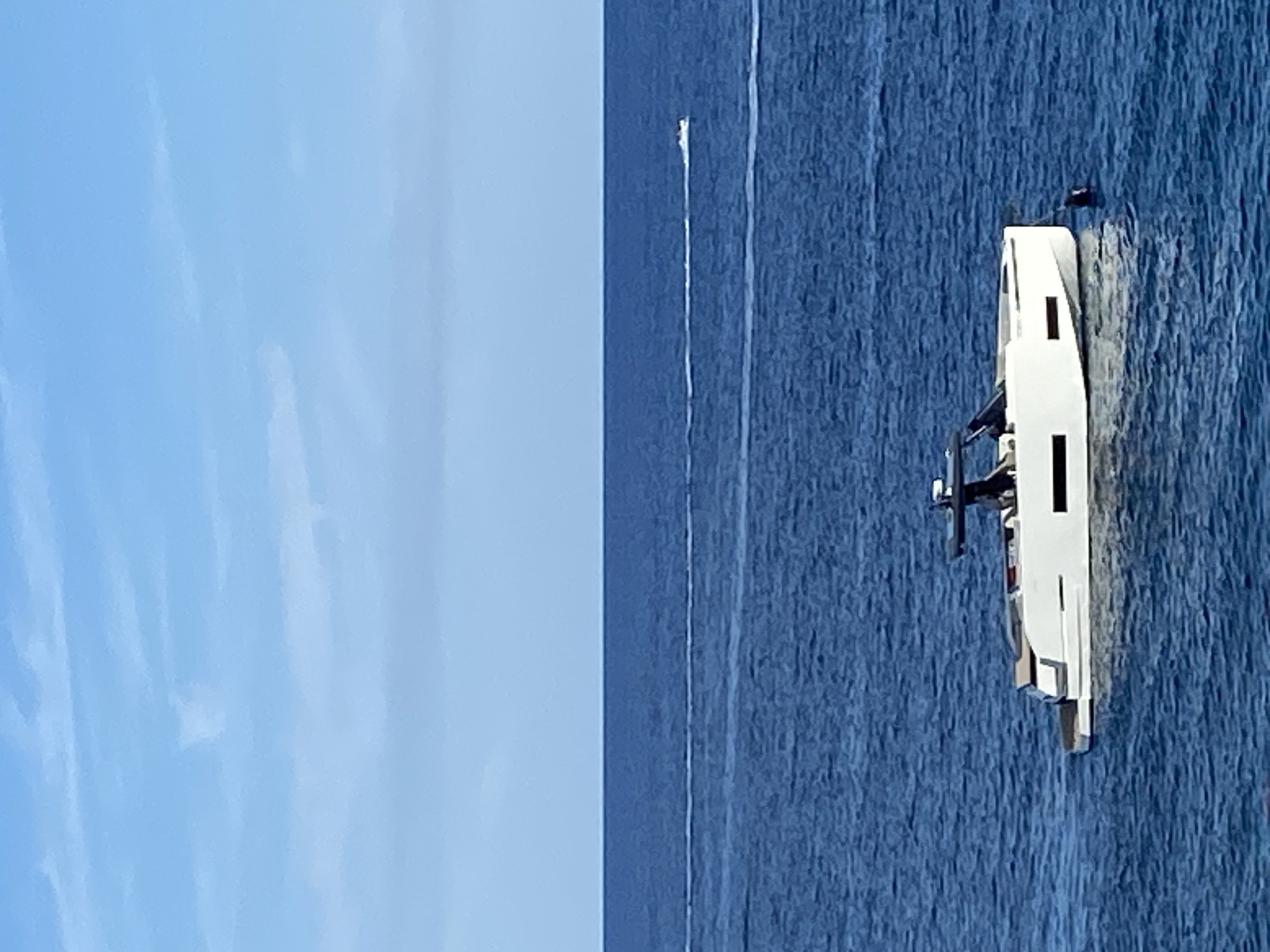 The image shows a clear blue sky above a calm, deep blue ocean. The horizon line is visible, separating the sky and sea. In the foreground, a modern white motor yacht is anchored. The yacht has a sleek design with dark-tinted windows and a prominent dark canopy or hardtop structure. A black buoy is visible in the water to the right of the yacht. In the middle distance, two smaller boats are visible, each leaving a white wake behind them, indicating they are moving across the water. The scene appears to be a tranquil day at sea, likely in a coastal area near Prijevor, Montenegro, given the context.