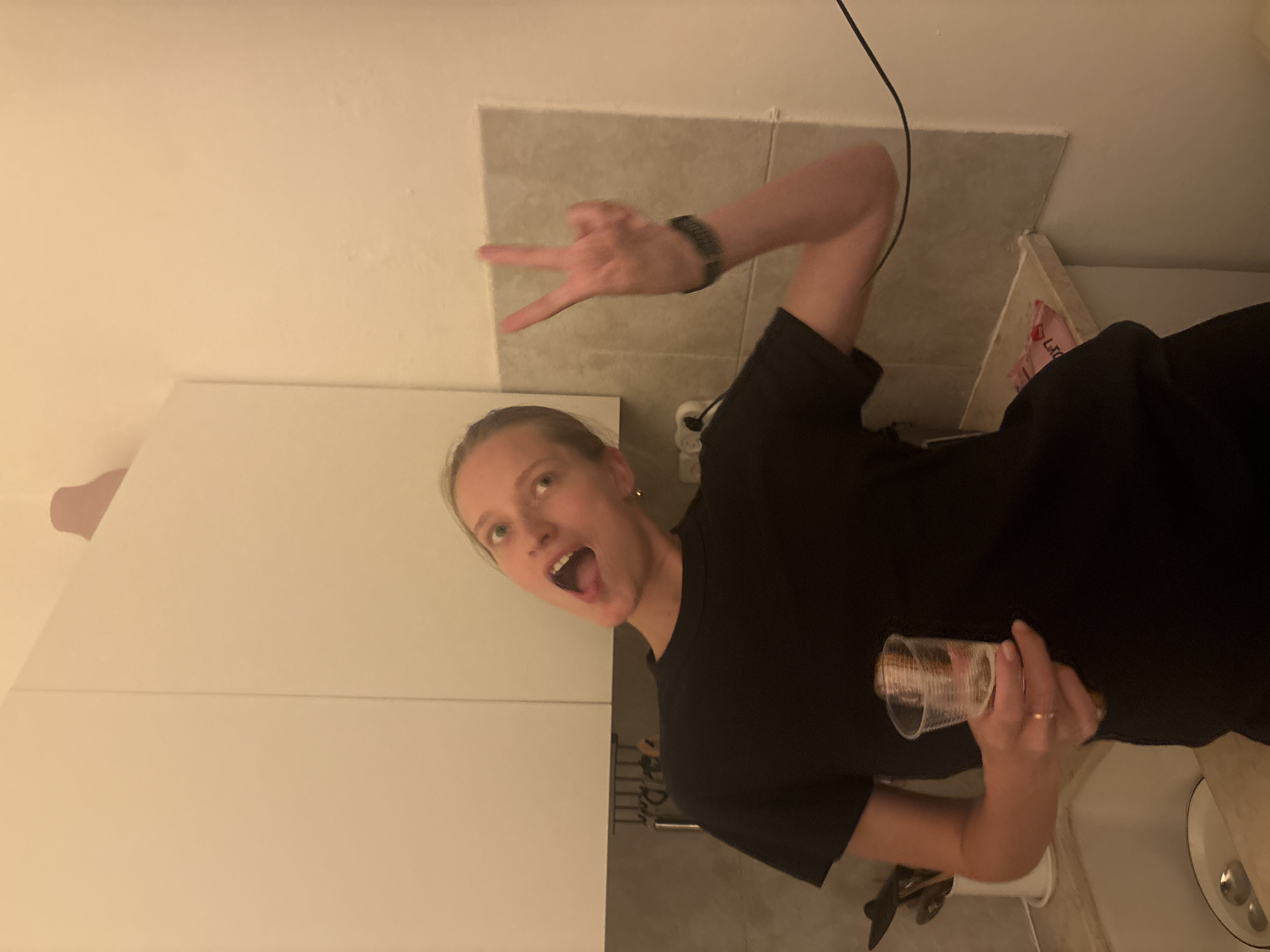 A woman with blonde hair pulled back is standing in what appears to be a kitchen. She is wearing a black t-shirt and has her mouth open, looking towards the camera. Her left hand is holding a plastic cup with a drink, and her right hand is raised, making a peace sign. She is wearing a watch on her right wrist and a ring on her left ring finger. Behind her, white kitchen cabinets are visible, along with a tiled backsplash and a sink. The scene is lit with warm lighting, suggesting an indoor setting. The presence of a sink and cabinets strongly indicates an indoor, domestic environment, likely a kitchen.
