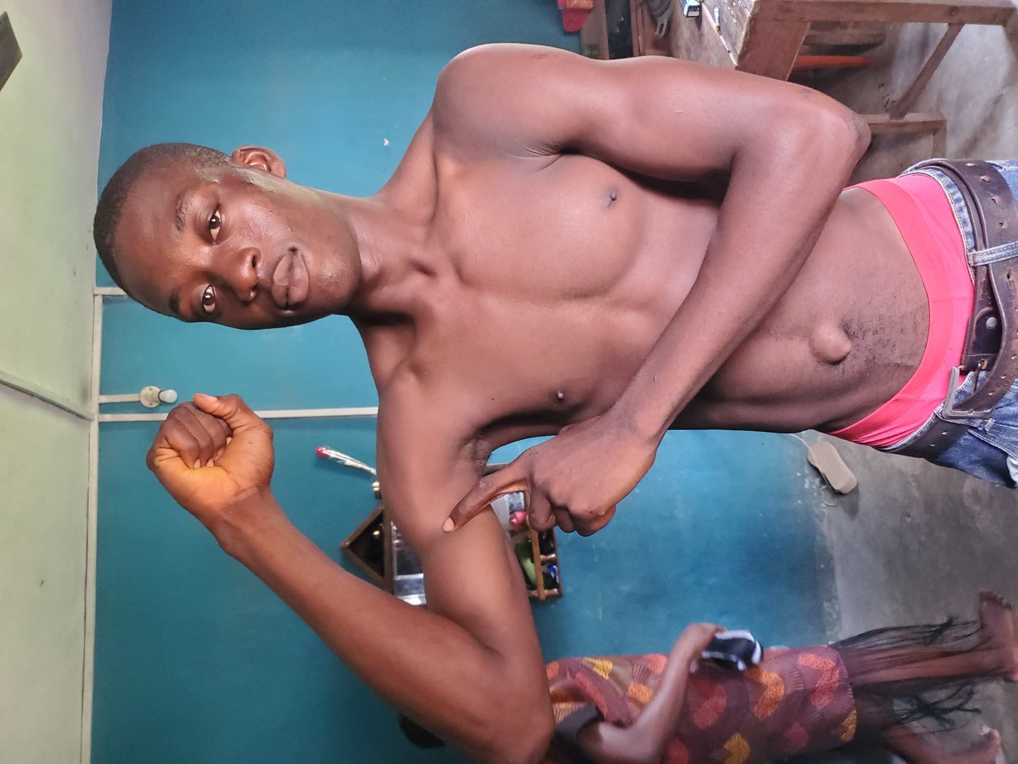 A shirtless dark-skinned man is indoors. He is wearing jeans with a brown belt and red underwear showing at the waist. He is flexing his left arm and pointing to his right arm. Another person is lying on the floor in the lower portion of the image, possibly asleep and covered. The location is Atani, Nigeria. The room has turquoise walls.