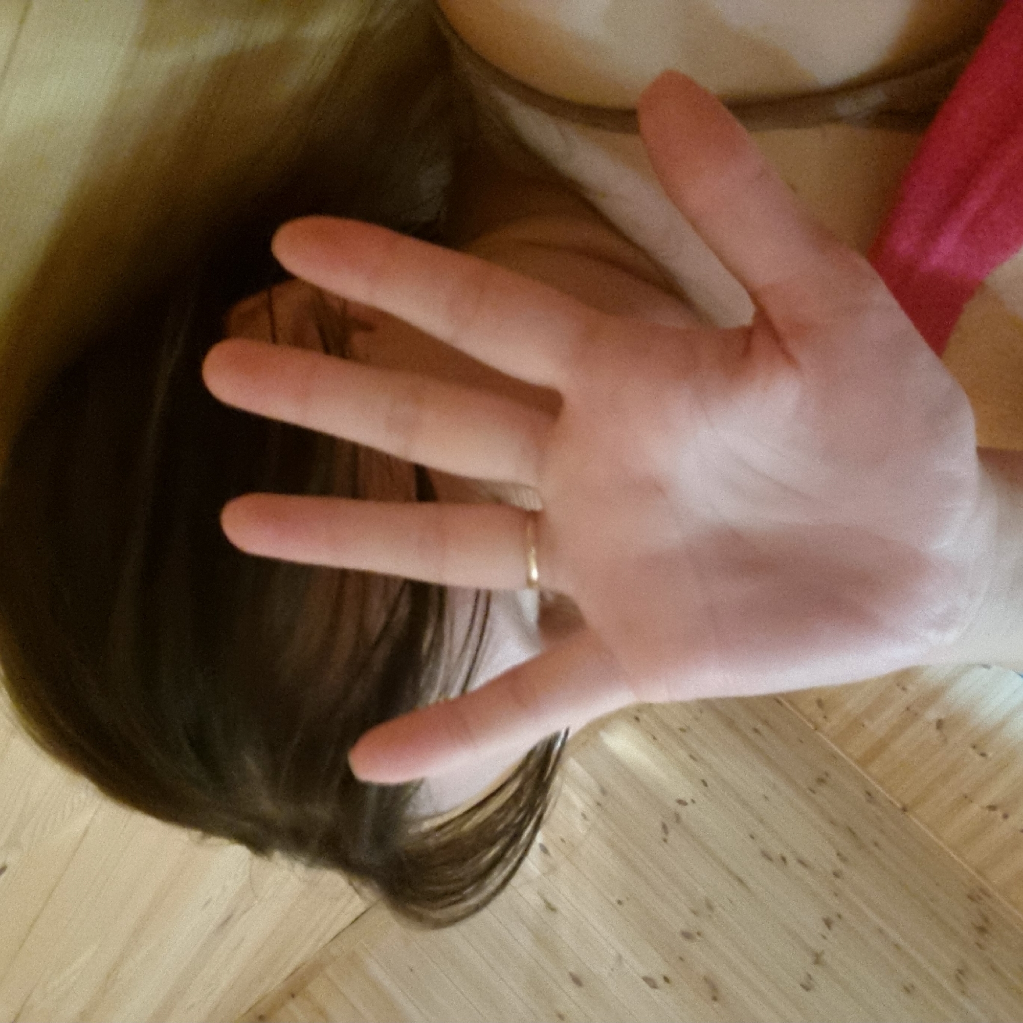 The image depicts a person's hand raised in front of their face, obscuring their features. The hand is positioned in the foreground, with fingers spread. A thin gold ring is visible on one of the fingers. The person's dark hair is visible to the left of the hand. The background appears to be a wooden floor. The scene is lit with artificial light, casting shadows. The provided location information, Dunnae-myeon, South Korea, is not visually represented in the image and cannot be confirmed from the visual content alone.