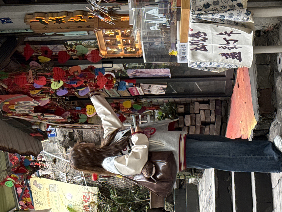 The image depicts a female individual engaging with a shop facade in a narrow, stepped alleyway, located in Yuzhong District, Chongqing, China. The subject, viewed in right profile, has long brown hair and is dressed in a light grey and off-white letterman-style jacket with red striped accents, wide-leg blue jeans, and white sneakers. She carries a dark brown hobo bag with a brown fluffy charm on her left shoulder and holds a small, light-colored object in her left hand. Her right arm is extended, and her hand touches one of the numerous multicolored, heart-shaped wooden ornaments, adorned with small bells and Chinese characters, which hang densely from the shop's exterior.
The shop entrance features a dark doorframe and a prominent, illuminated wooden sign with vertical Chinese characters, including "山城" (Shancheng), mounted to its right. Below the hanging ornaments, a display table covered with a white, blue-patterned tablecloth holds a clear acrylic case containing miniature city models. A vertical banner on the table's front displays "山城 盖戳卡" (Shancheng Stamp Card). A smaller, lighted display unit with individual compartments is visible behind the table. The ground immediately in front of the shop entrance is a reddish-orange painted section, contrasting with the surrounding irregular stone steps and paved surfaces. The background reveals exposed brick and stone walls, potted greenery to the left, and multi-story structures climbing uphill, characteristic of Chongqing's hilly urban environment. Utility lines are visible overhead. A blue street sign marked "1-1" is affixed above the shop's door.