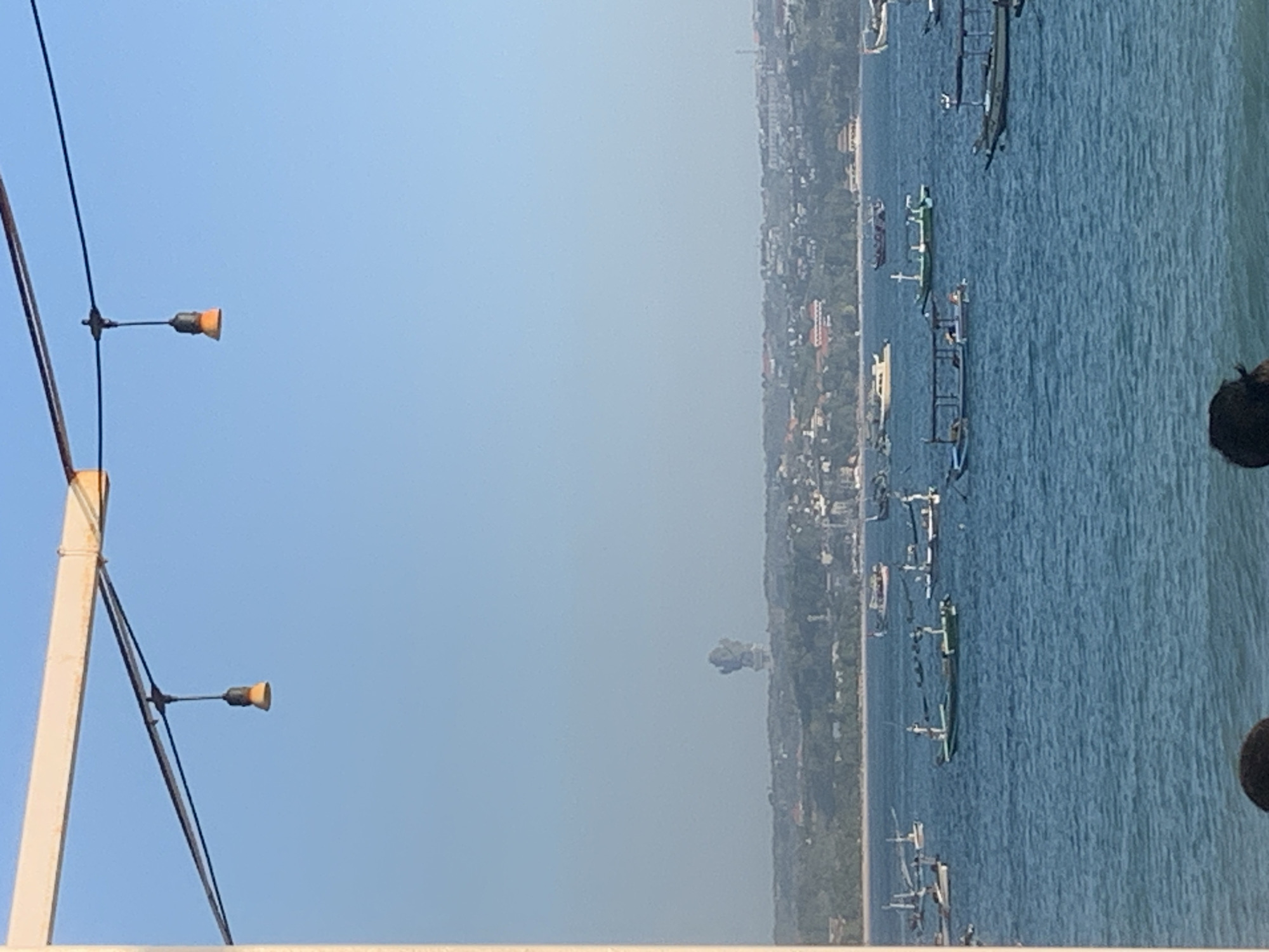 The image shows a wide, clear blue sky with a hint of haze. In the foreground, a white pole with two light fixtures is visible on the left side, attached to black wires. The light fixtures are amber-colored and extend outwards.
The background depicts a coastal scene. A shoreline with trees and buildings runs along the edge of a calm, blue body of water. Several long, narrow boats are anchored in the water, appearing to be traditional fishing vessels. The boats are clustered together in some areas and more spread out in others. In the distance, a hazy, dark object is visible on the land, possibly a structure or a rock formation. The lighting suggests it is daytime, likely morning or afternoon, with a bright sky. The overall atmosphere is serene and tranquil, hinting at a coastal town or village in Denpasar, Indonesia.