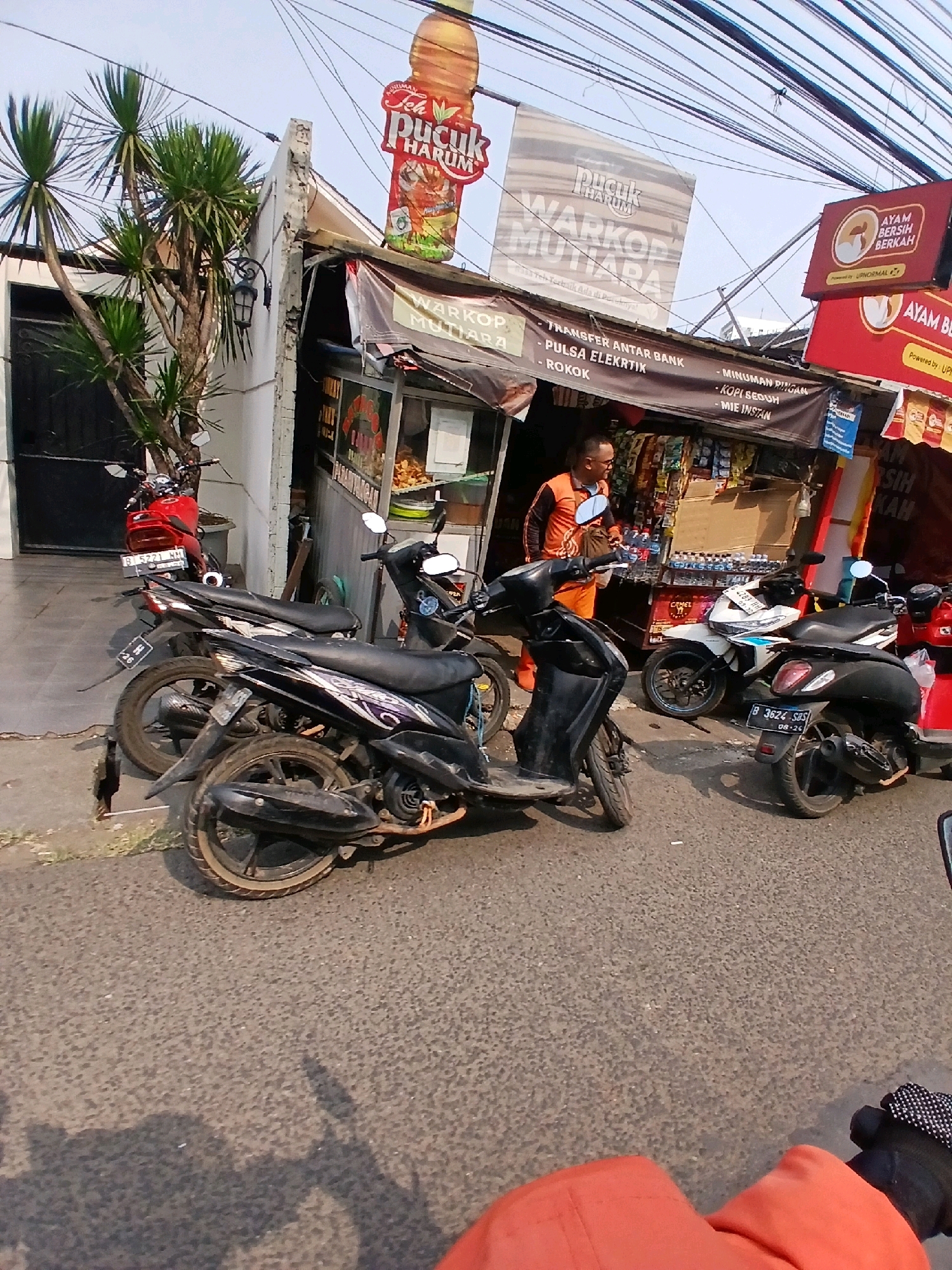 This image depicts a street scene in South Jakarta, Indonesia, featuring a small storefront or kiosk. The kiosk appears to be a warung, selling various items such as drinks, instant noodles, and cigarettes, indicated by signage that reads "WARKOP MUTIARA." A man in an orange shirt is visible behind the counter. Several motorcycles are parked in front of the establishment, including a red one and multiple dark-colored scooters. A prominent advertisement for "Teh Pucuk Harum" is visible overhead, along with other advertisements for "AYAM BERSIH BERKAH." The environment is characterized by overhead electrical wires, typical of urban areas in Indonesia. The time of day appears to be daytime, with sunlight casting shadows.