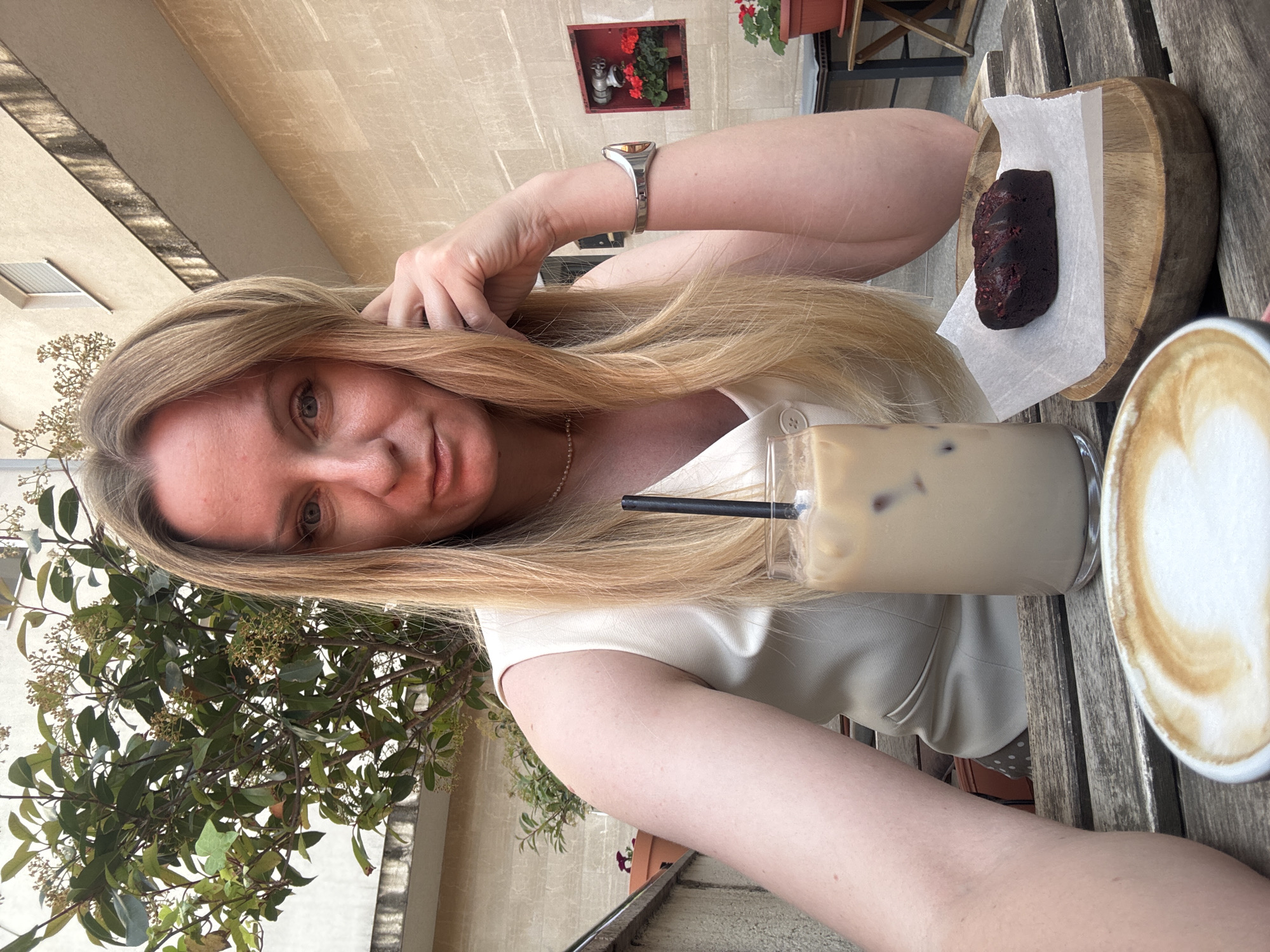 A close-up selfie captures a woman with long, straight blonde hair and light-colored eyes, looking directly at the camera with a calm expression. She is wearing a light-colored top, possibly cream or off-white, and a delicate necklace. A silver-toned watch adorns her left wrist.
She is seated at a rustic wooden table, where several items are laid out. To her right, there's a glass of iced coffee with a black straw. Next to it, a white cup containing a hot coffee, likely a latte or cappuccino, displays visible foam art. A dark, oblong pastry, possibly a chocolate brownie or cake, rests on a piece of white parchment paper within a small wooden serving tray.
The setting appears to be an outdoor café or restaurant in Budva, Montenegro. The background features light-colored building walls, suggesting a warm climate. To the left, lush green foliage with small white or light-colored blooms or berries is visible. In the upper right corner, a red wall-mounted planter with vibrant red geraniums adds a pop of color against the beige wall. The lighting indicates it is daytime, likely a pleasant morning or afternoon. No text is visible in the image.