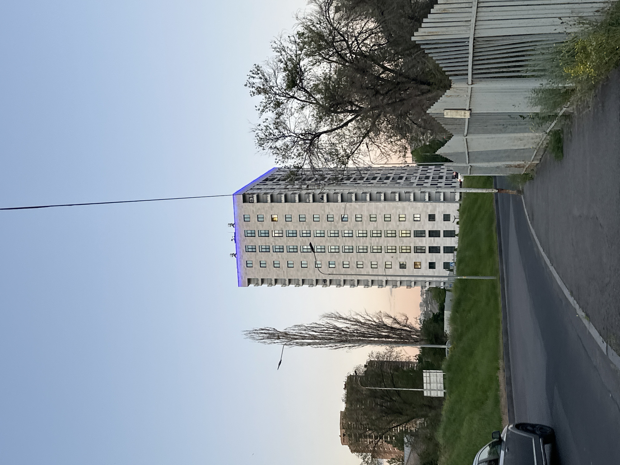 A tall, multi-story building with a repetitive pattern of windows and balconies stands adjacent to a road and green space. The building appears to be constructed of light-colored concrete or stone and has a line of blue lights at the top. To the right of the building, a section of a fence or barrier is visible, partially obscured by trees. A road runs alongside the green space, with a dark-colored vehicle partially visible in the lower right corner. The sky is a light blue color, indicating either morning or evening light. The presence of specific architectural styles and the overall urban environment suggest that the location is likely Yerevan, Armenia.