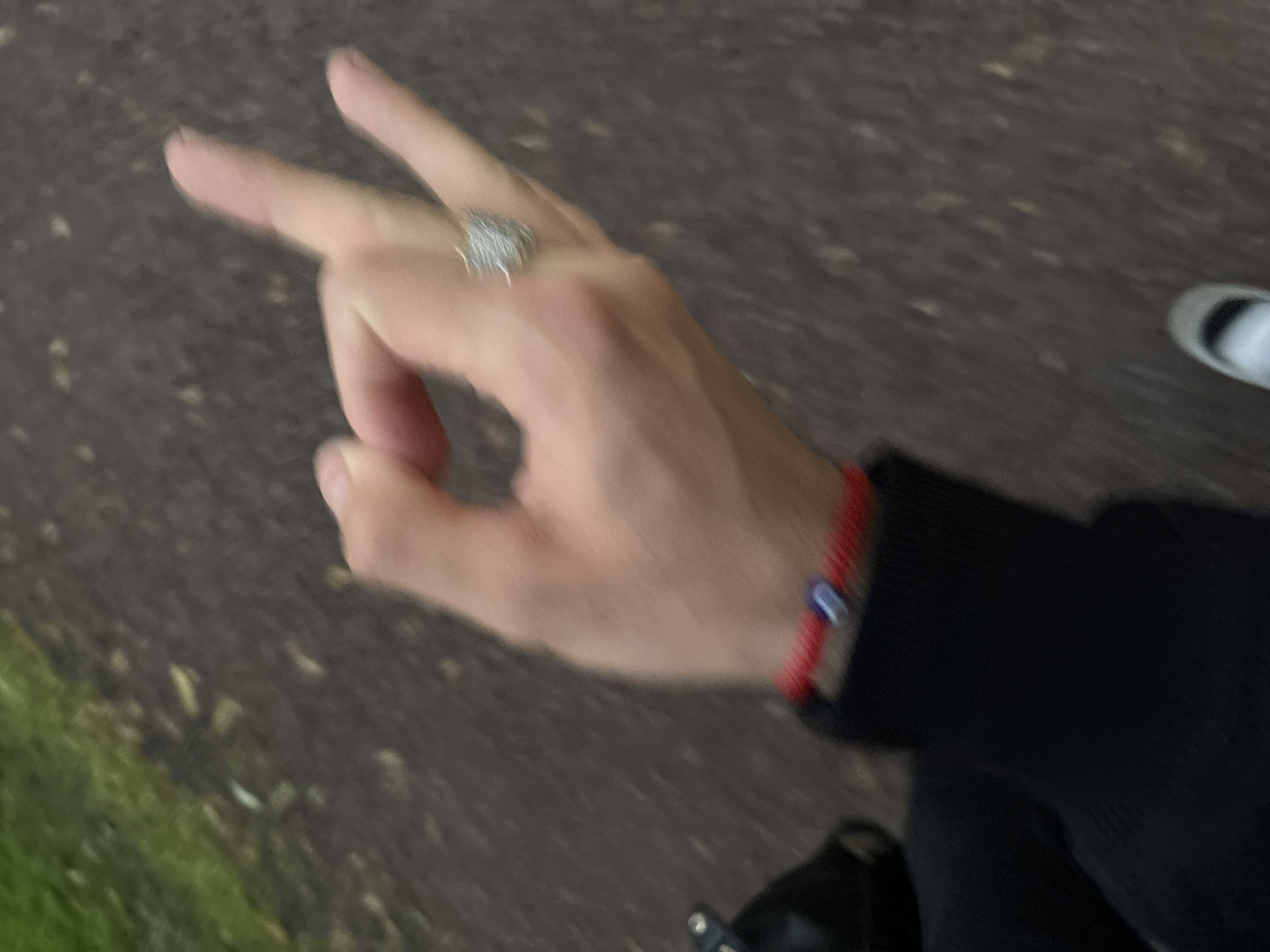 A first-person perspective shot shows a person's left hand and wrist. The hand is in the foreground, with the thumb and index finger forming a circle, and the middle and ring fingers extended upwards. The person is wearing a ring with a spiky, metallic design on the middle finger and a red beaded bracelet with a blue evil eye charm on the wrist. The person is wearing a black long-sleeved garment, partially visible on the left. The background is blurred, suggesting movement. It appears to be an outdoor setting with a paved path and some grass on the left. The ground is a dark, mottled grey surface, possibly asphalt or gravel. The lighting is overcast. Based on the provided crop hinting at a location in Pori, Finland, it is plausible this scene is in a park or walking path within the city. The image captures a brief moment in time, perhaps while walking or standing still, with the focus on the hand and its adornments against a softly blurred natural and urban backdrop.