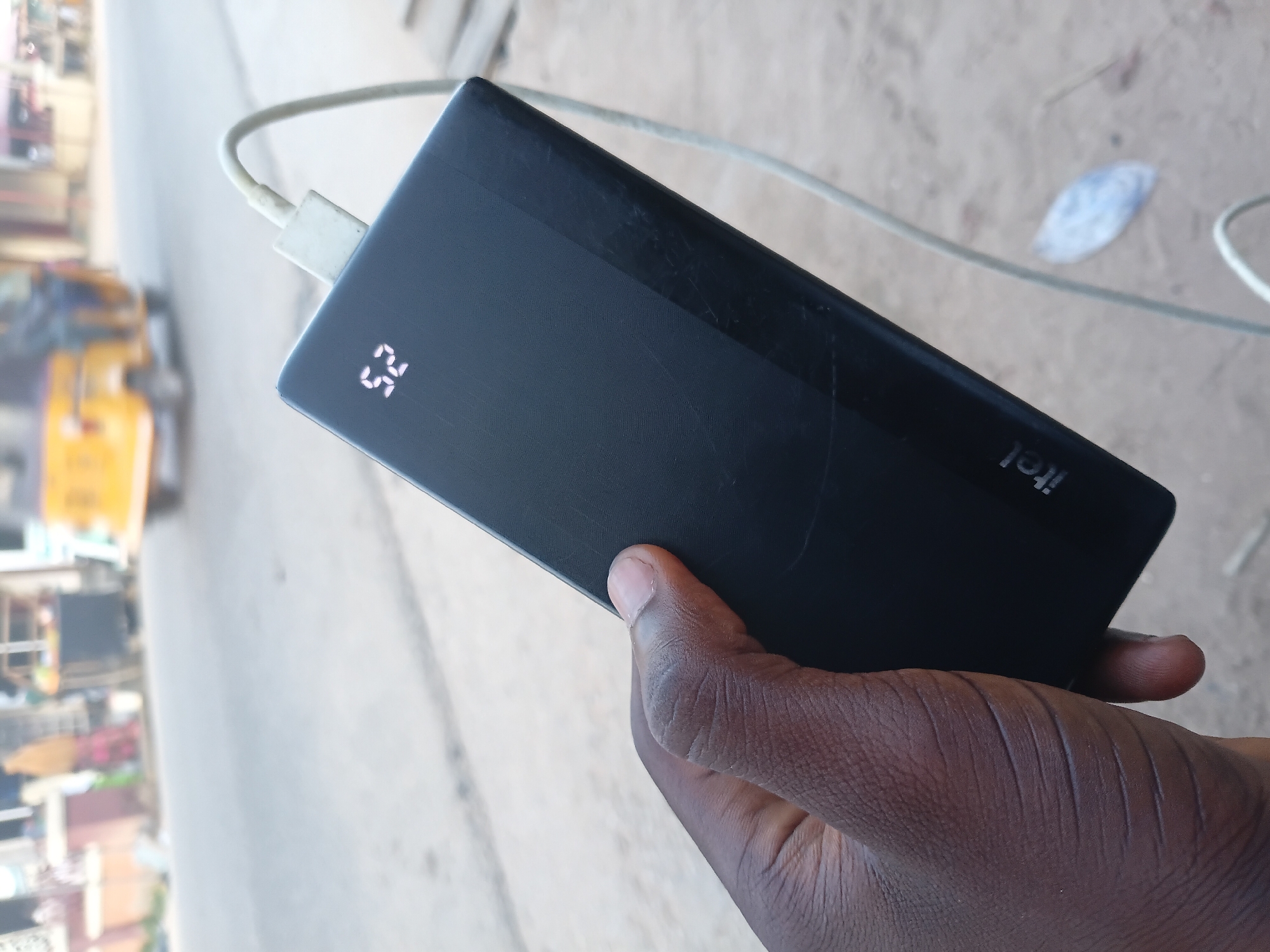 A black power bank with a white charging cable attached is held in a dark-skinned person's hand. The power bank displays "52%" on its digital screen. The background is blurry and appears to be an outdoor street scene in the city of Jalingo, Nigeria, with a yellow three-wheeled vehicle and buildings visible. The ground is dirt or pavement.