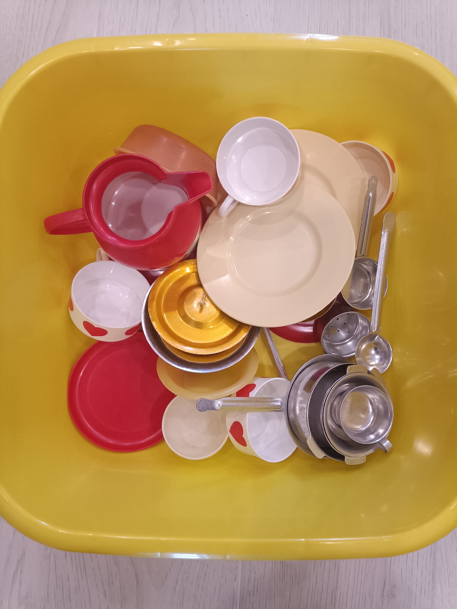 A yellow plastic basin is filled with assorted children's toy dishes. The dishes include a red pitcher, several white and red-patterned cups, a yellow plate, and several silver metal ladles and strainers. The basin is placed on a light-colored wooden surface. There are no people or animals visible in the image. The scene is domestic, and there is no information to suggest it is located in Temirtau, Kazakhstan.