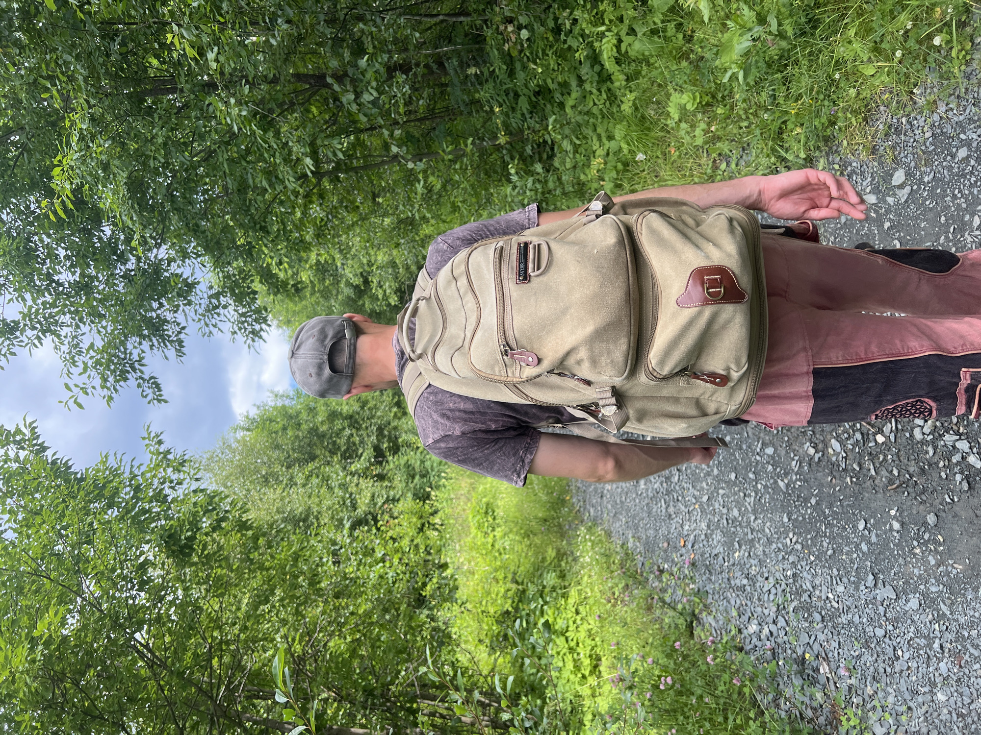 A person is walking away from the camera on a gravel path in a forest. The person is wearing a grey baseball cap, a dark t-shirt, and pink and black pants. They are carrying a large, tan backpack. The path is lined with green trees and grass. The sky is partly cloudy. The location is the city of Gubakha, Russia.