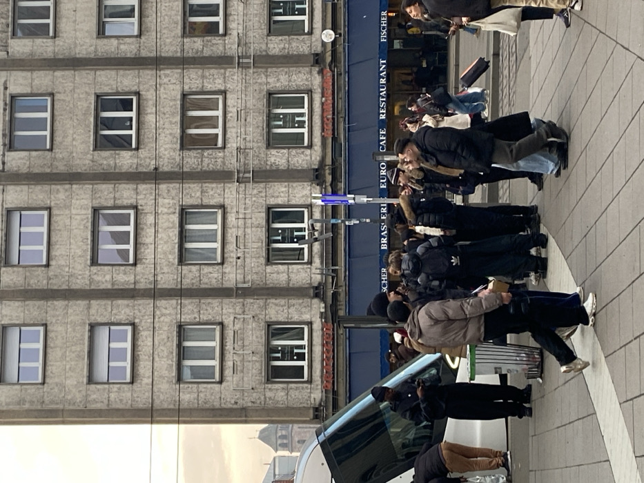 This media file captures an urban street scene in Strasbourg, France, showing a crowd of people on a sidewalk next to a building. The multi-story building features a light-colored, textured facade with numerous rectangular windows across its many floors.
At street level, the building houses an establishment with a prominent blue awning and signage that reads "EURO CAFE RESTAURANT BRASSERIE FISCHER." A diverse group of pedestrians, dressed in winter or cool-weather clothing such as jackets, coats, and long pants, are visible on the sidewalk. Some individuals appear to be walking, while others are standing and observing, suggesting a gathering or typical street activity.
Notable details include a light pole with glowing blue elements in the mid-ground. In the foreground, part of a white vehicle, possibly a van or car, is visible with its door open, and a person is bending over near it. The lighting suggests it is daytime, possibly overcast or late afternoon. A "ADIDAS" logo is discernible on a backpack worn by one of the individuals in the crowd. The paved sidewalk features light-colored circular patterns. The overall scene depicts a lively city environment.