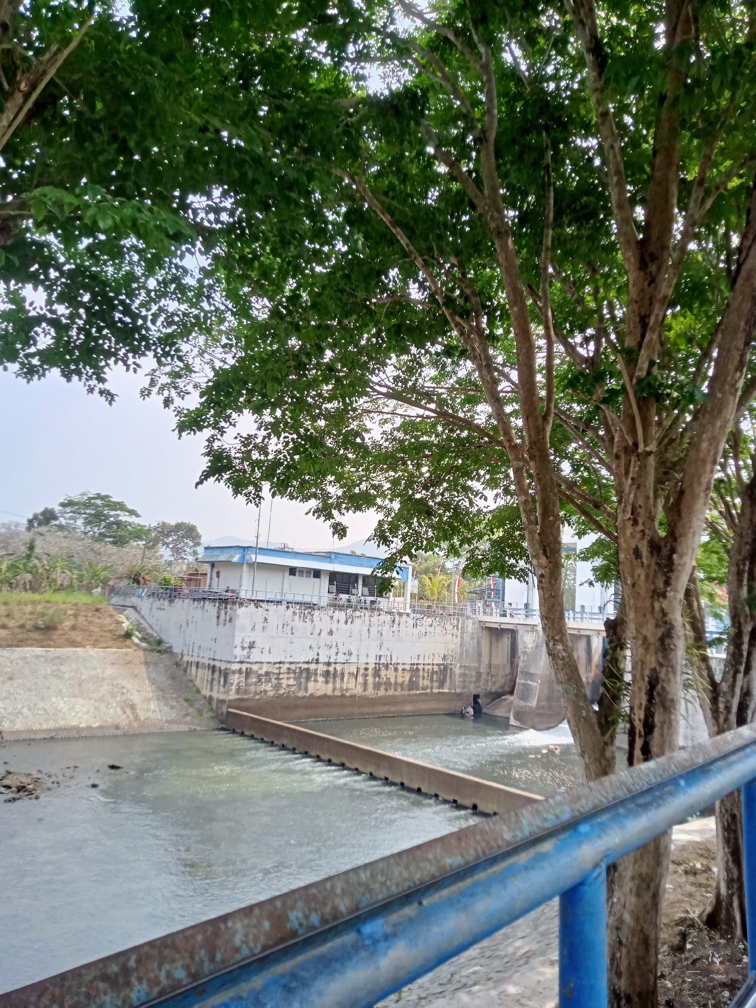 A full shot captures a dam structure with a building on top, situated in the city of Tiudan, Indonesia. The dam, constructed from white concrete, features a series of gates that are partially open, allowing water to flow through. Adjacent to the dam is a grey, sloped embankment, and beyond that, a sandy area with some vegetation. In the foreground, a blue metal railing runs across the frame, partially obscuring the view of the dam and its surroundings. Several trees with green foliage are visible, with their branches extending into the frame from the right and top. The sky above is overcast.