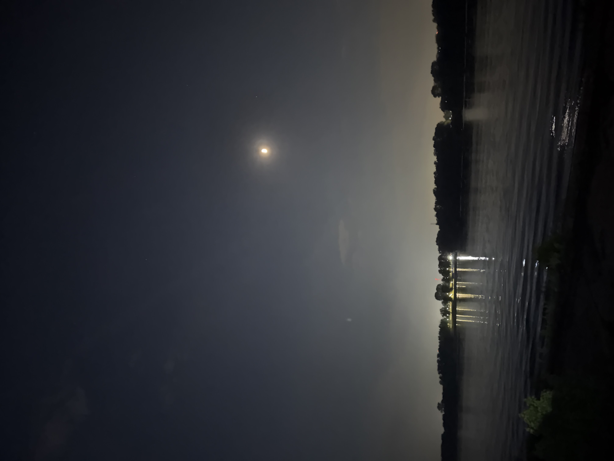 The image depicts a dark, nocturnal scene with a body of water, possibly a lake or river, under a dim moonlit sky. The moon is visible as a small, bright orb in the upper left quadrant of the image. The water's surface reflects some light, creating a shimmering effect. The opposite shoreline is lined with trees, appearing as a dark, undefined mass. Some artificial lights are visible along the shoreline, reflecting in the water. The overall mood is quiet and serene. Based on the provided information that it is located in the city of Nizhny Novgorod, Russia, the body of water is likely the Volga or Oka river, or one of the lakes within the city limits.