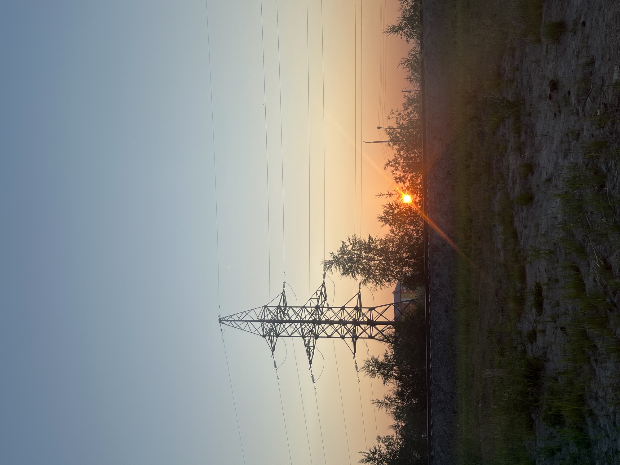 This is a landscape photograph taken in Novy Urengoy, Russia, oriented vertically. The scene depicts a sunset with a soft gradient of blue transitioning to a warm orange glow in the sky. Several power lines are visible, stretching horizontally across the frame. In the center, a tall, lattice-style transmission tower is silhouetted against the sky.
To the right of the tower, trees with sparse foliage are present, and the setting sun is a bright, distinct orb peeking through the branches, casting a lens flare. Further to the right, a rough, unpaved ground surface with scattered grass and some debris is visible, suggesting an outdoor, possibly rural or undeveloped area. A railway track runs along the edge of this ground. In the background, beyond the trees and power lines, a building with an orange roof and yellow walls is partially visible. The overall mood is serene and tranquil, characteristic of a quiet evening. The time of day is clearly sunset, indicated by the warm, low-angled sunlight and the colors in the sky. There are no people visible in the image, and no text is present.