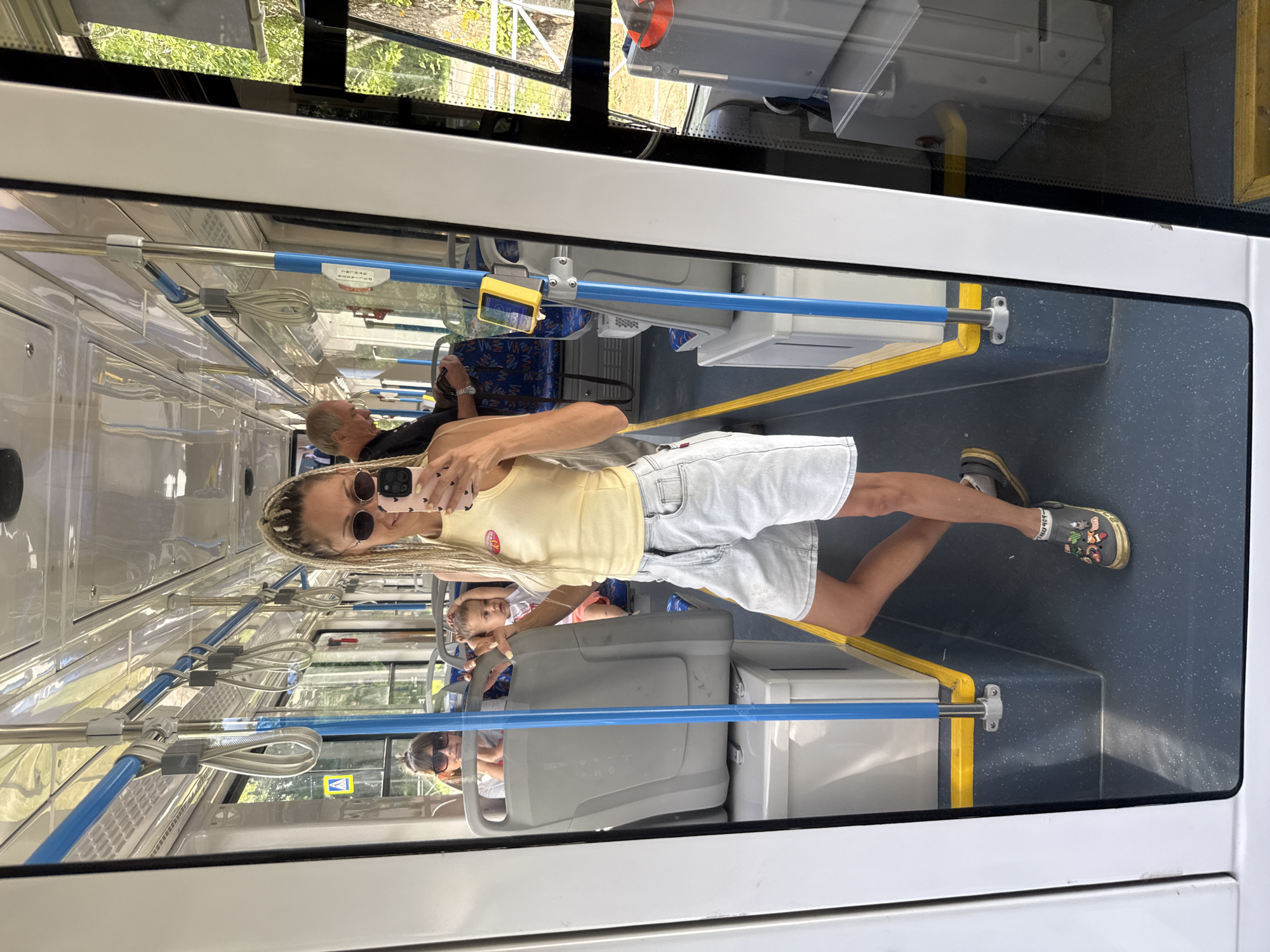 The image is a mirror reflection taken inside a public transport vehicle, likely a bus or tram, on a sunny day. The reflection shows a woman with blonde braided hair, wearing sunglasses, a yellow tank top, and light-wash denim shorts. She is holding a pink smartphone and taking a selfie. In the background, other passengers are visible, including a woman with sunglasses and a child in a seat behind her. Another man is seated further back. The interior of the vehicle features blue and yellow grab bars, gray seating, and blue patterned upholstery. Outside the windows, trees and foliage suggest a green, possibly suburban or rural, environment. The scene appears to be taking place in Rusanovo, Russia, though this location is not visually confirmed within the image itself.