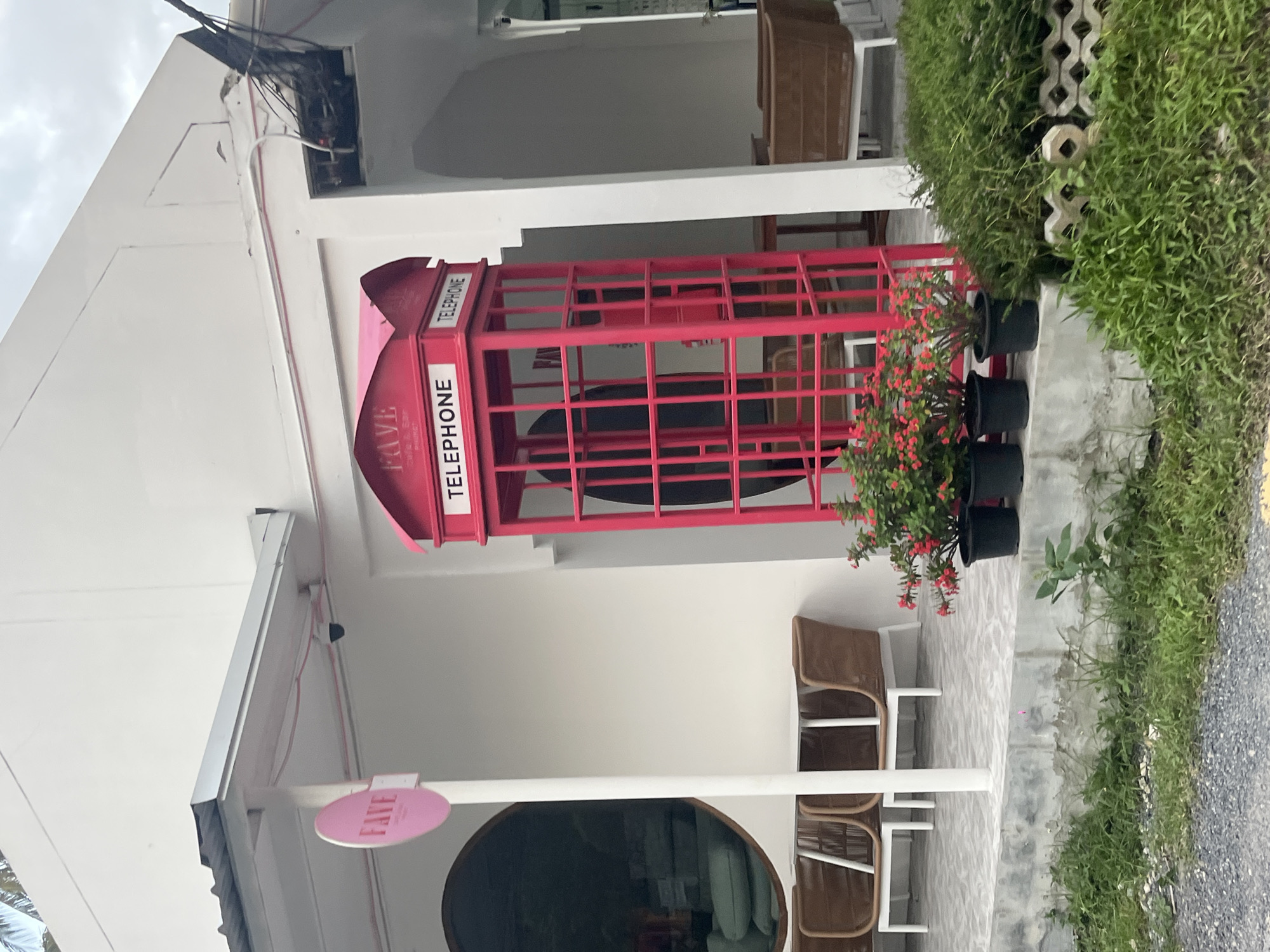 The image shows a cafe with a prominent red telephone booth as a decorative object. The telephone booth is placed near the entrance of the cafe, which is a white building with a covered outdoor seating area. There are several potted plants, including one with small red flowers, arranged around the telephone booth. The cafe has outdoor seating with chairs and tables, some made of rattan. In the background, there is a wall with textured concrete blocks and lush green grass. The scene appears to be in a tropical or subtropical location, suggested by the vegetation. There is no activity taking place, and the image seems to capture a static view of the cafe's exterior. The lighting suggests it could be daytime, possibly overcast. The visible text includes "TELEPHONE" written on the telephone booth and "FAVE" on a pink oval sign near the entrance. The overall impression is a visually interesting and welcoming cafe entrance.