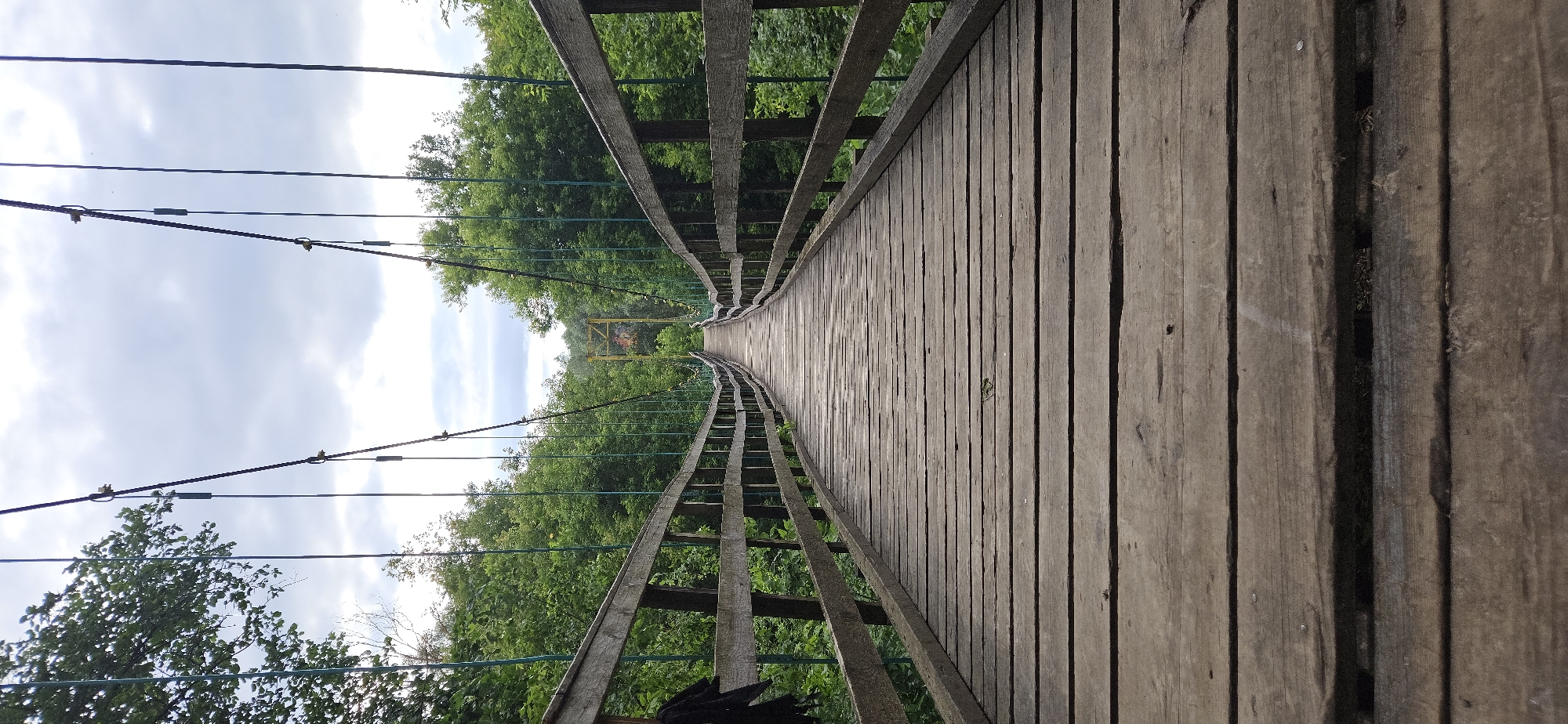 The image depicts a wooden suspension bridge with a high perspective, looking down the length of the walkway. The bridge is surrounded by lush green trees, suggesting a natural or park-like setting. The sky above is partly cloudy, indicating daytime. The bridge itself is constructed with wooden planks for the walkway and wooden railings on either side, supported by cables. In the distance, a yellow structure is visible, possibly part of a larger attraction or another bridge. The scene appears to be a peaceful and serene environment, ideal for a leisurely walk or exploration. There is no visible text in the image.