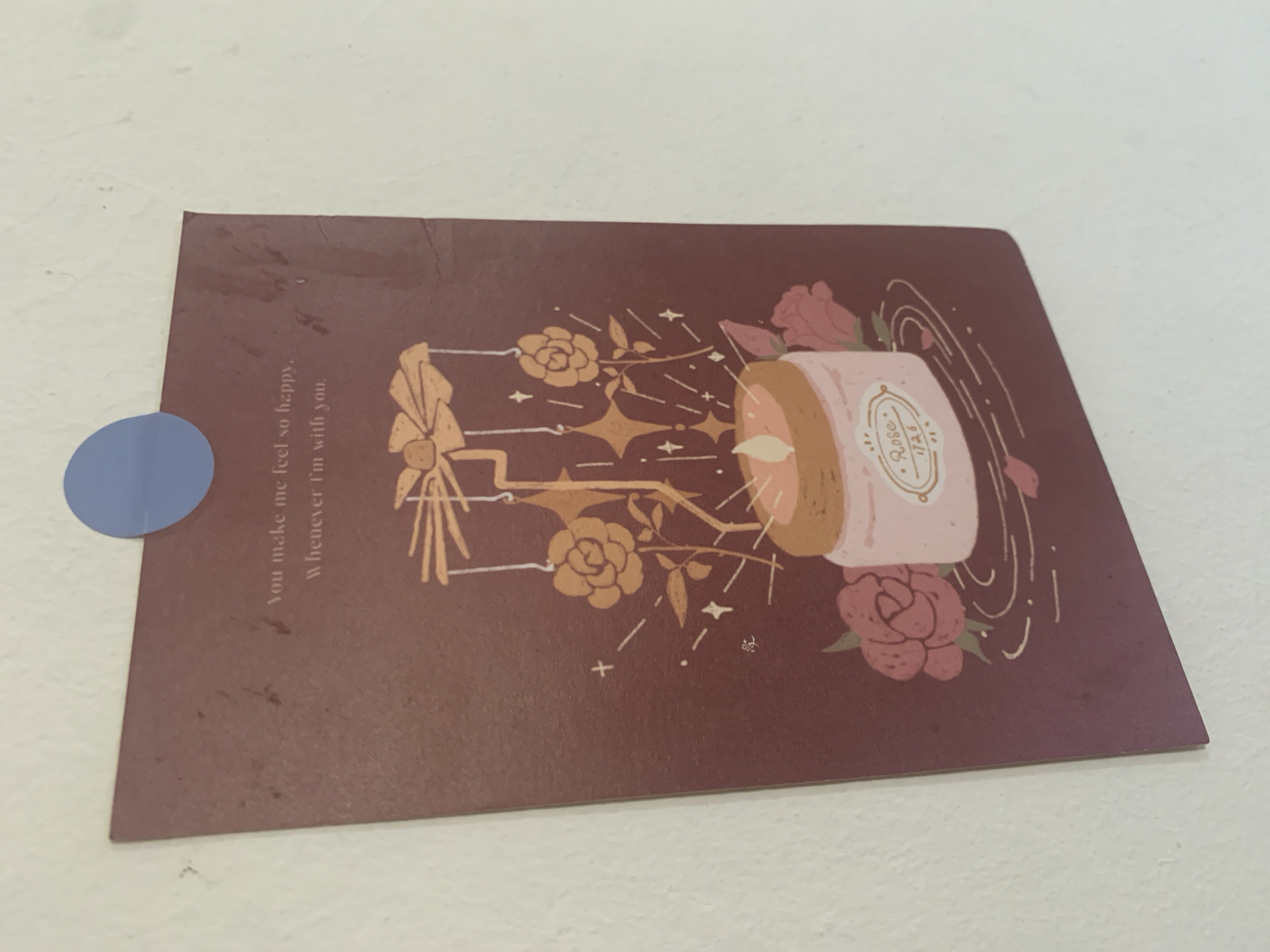 A reddish-brown postcard is attached to a white wall with a blue circular sticker in the upper right corner. The postcard features an illustration of a candle in a pink jar, surrounded by roses and decorative elements. Text above the illustration reads, "You make me feel so happy. Whenever I'm with you." The postcard is positioned at an angle, with the top edge angled downwards from left to right. The wall appears textured and slightly grimy. This scene is located in Kuala Pembuang, Indonesia.
