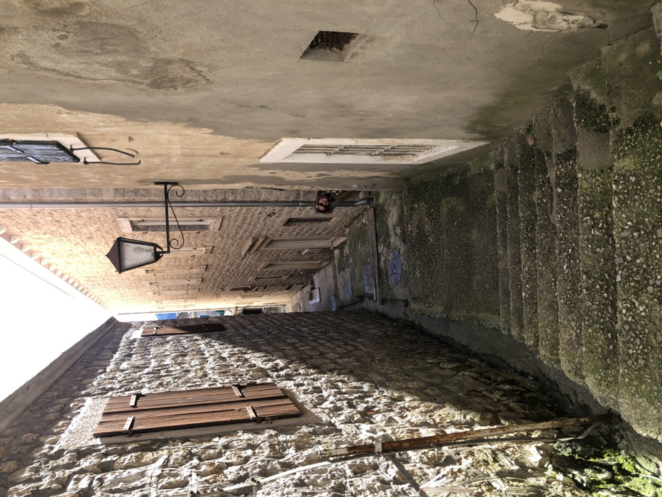 The image displays a narrow, stepped alleyway in Herceg Novi, Montenegro, descending into the distance under strong daylight.
On the left, a building constructed of light-colored, rough-hewn stone blocks features two visible windows with closed, dark brown wooden shutters. A rusty metal pipe runs vertically along the lower portion of this wall. The wall is primarily in shadow.
On the right, a building with a light beige, plastered facade shows signs of age, including peeling plaster and vertical streaks of discoloration. An ornate black metal street lamp with a glass lantern is affixed to this wall. Below the lamp, a vertical metal downspout is visible. Multiple windows are present, with one in the lower foreground featuring a white frame and metal grates. A small, square opening is also visible on this wall.
The alley's surface consists of worn, grey concrete steps in the foreground, transitioning to a flatter, paved section further back. Both the steps and pavement exhibit patches of green moss or discoloration. Several circular utility access covers are embedded in the pavement in the mid-ground. A narrow drainage channel runs along the base of the left wall.
In the mid-ground, a person is partially visible, peeking from behind the corner of the building on the right. The individual's head and upper torso are discernible, revealing long, dark hair and a patterned garment. The person is looking towards the viewer's position.