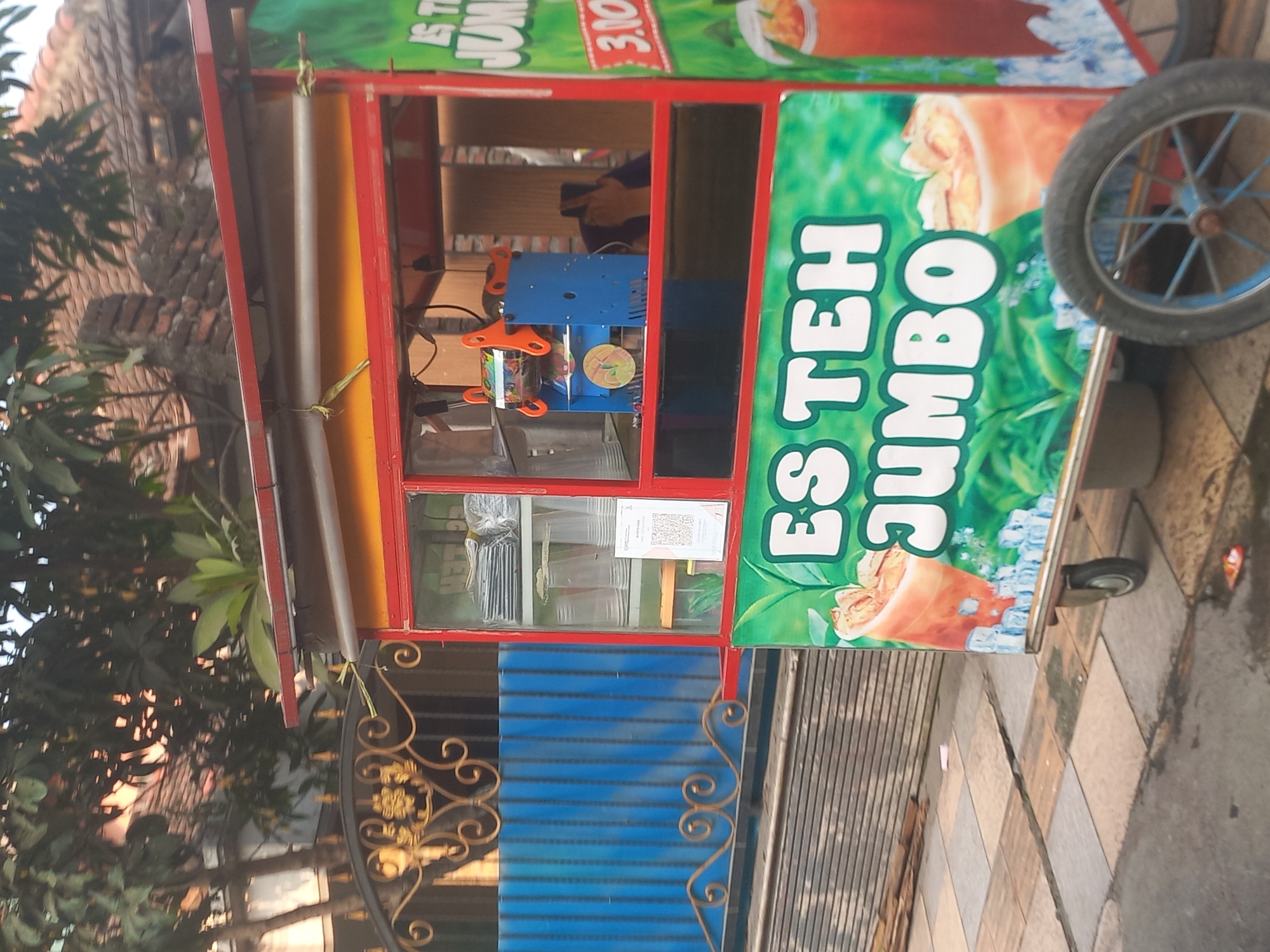 An outdoor shot captures a food cart selling iced tea in Jombang, Indonesia. The cart, painted red and yellow, features a clear display case containing plastic cups and other items. A machine for sealing cups is visible inside. The cart's side displays large white text in Indonesian that reads "Es Teh Jumbo," meaning "Jumbo Iced Tea," with an illustration of iced tea and ice cubes. The cart has wheels and is parked on a paved area. In the background, a blue metal fence with decorative elements and a glimpse of a building with a tiled roof are visible. Trees with dense foliage overhang the scene. The lighting suggests it is daytime.