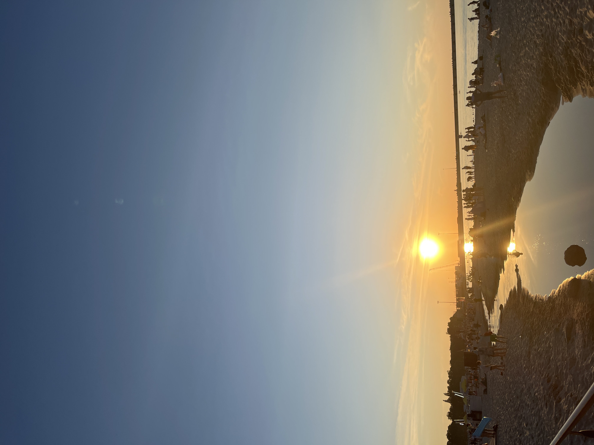 The image depicts a beach scene at sunset in Pionersky, Russia. The sun is low on the horizon, casting a warm orange glow across the sky and reflecting on the calm water of a shallow stream on the sand. Numerous people are visible on the beach, some standing, others sitting or lying down. They appear to be enjoying the evening, with some walking along the shore or interacting with others. In the background, a body of water, likely the Baltic Sea, stretches to the horizon, with a faint silhouette of structures and cranes on the far side. The beach itself is sandy, with some scattered debris or rocks. The sky transitions from a bright orange near the sun to a soft blue higher up.