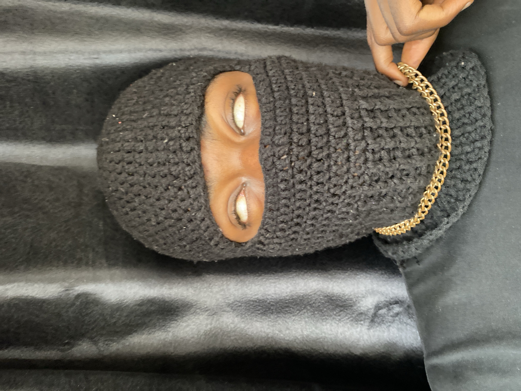 A person wearing a black crocheted balaclava lies on a dark, striated surface. The balaclava has two eye openings. A hand holds a gold chain near the right side of the balaclava. The setting appears to be indoors. No specific details indicate the location is in Barakin Akawo, Nigeria.