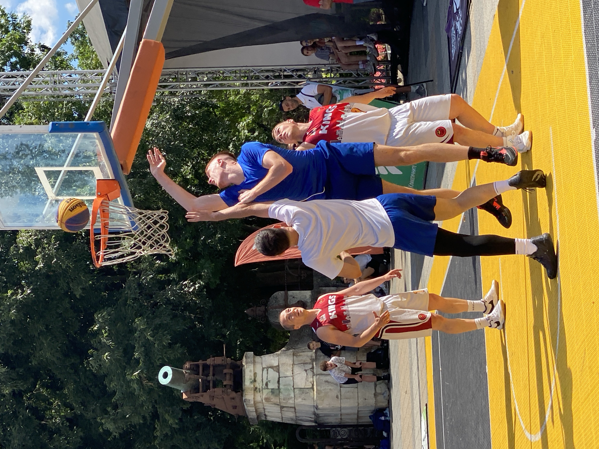 In Pleven, Bulgaria, an outdoor basketball game is underway on a yellow and grey court. Four young men, dressed in basketball attire, are actively engaged in play near a basketball hoop. One player, wearing a blue jersey and shorts, is in mid-air, reaching for the ball with his right hand as it nears the hoop. Two other players, one in a white jersey with red trim and white shorts, and another in a white t-shirt and blue shorts, are positioned beneath the hoop, looking upwards at the ball. A fourth player, also in a white jersey with red trim and white shorts, stands nearby, observing the action. In the background, a historic cannon monument is visible, surrounded by trees. Spectators can be seen seated and standing in the background, watching the game. The sky is partly cloudy.