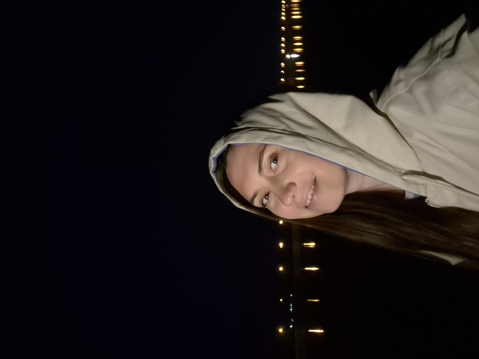 A low-angle, medium shot shows a woman wearing a hooded jacket and smiling, against a dark background punctuated by vertical lines of yellow lights. The woman has light skin, dark hair, and is positioned off-center towards the right, with her head slightly tilted. She appears to be wearing a jacket, the hood framing her face. The dark background has scattered yellow lights arranged in vertical columns, suggesting city lights reflected in water or from a bridge at night. The location is Unknown, Unknown, as the image does not contain identifiable landmarks or contextual information to determine its exact geographic location within a city.